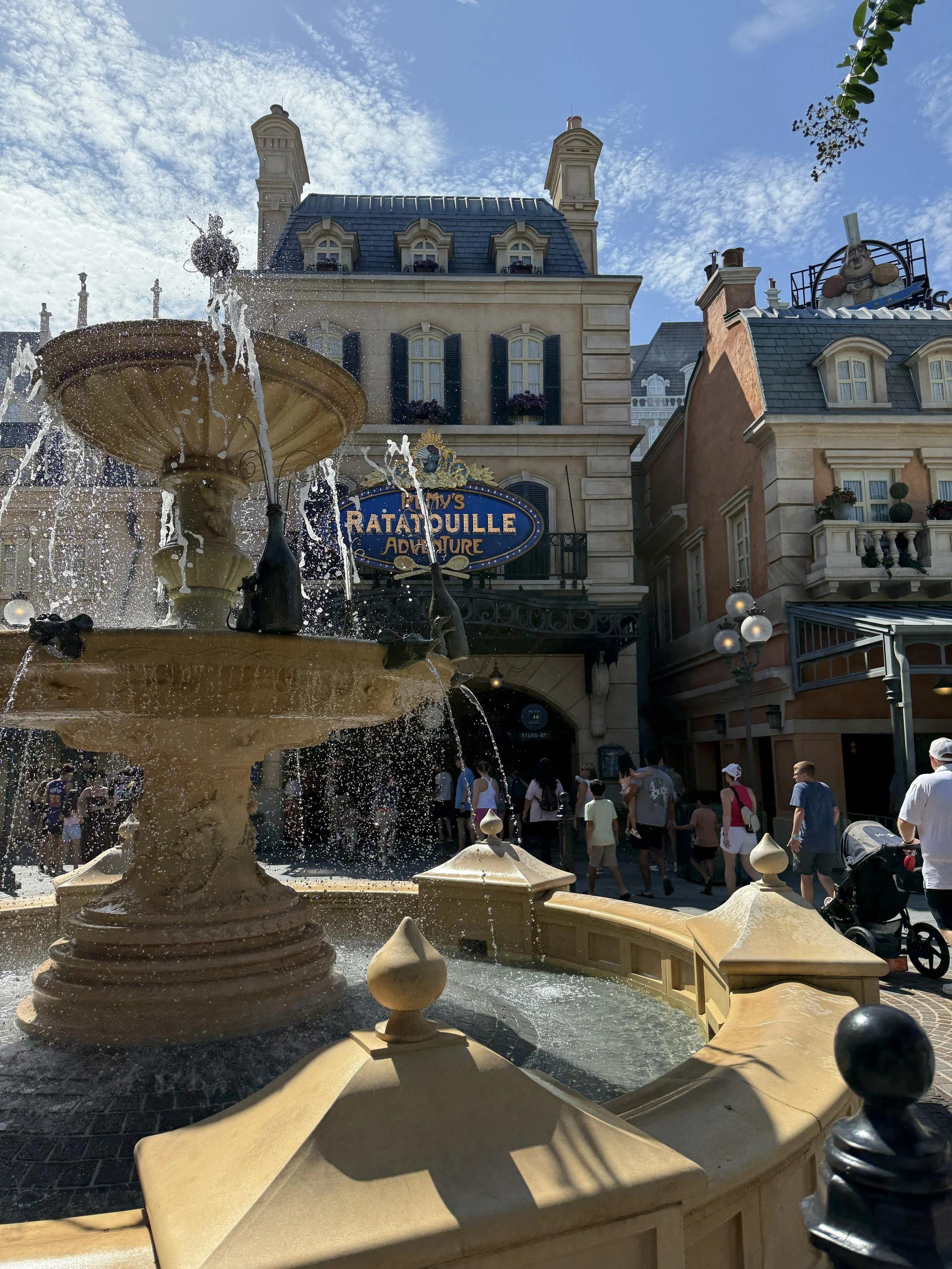 A castle-like building with a sign that reads 'Ratatouille Adventure' and a decorative fountain in the foreground, with people walking below and a partly cloudy sky above.