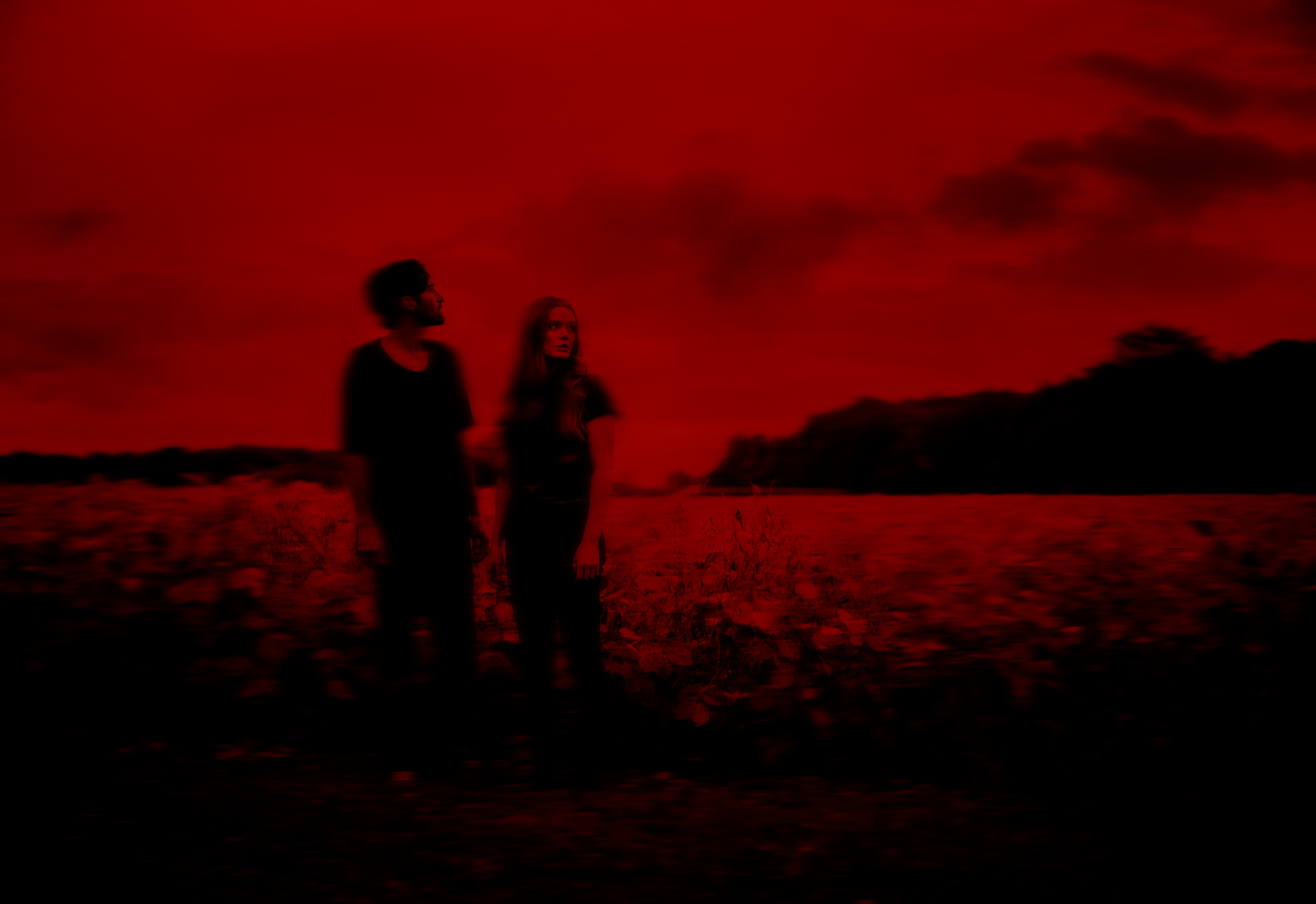 Silhouettes of a man and woman standing outdoors against a dark red sky with clouds.