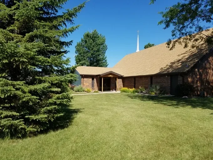 A brick church with a brown roof, surrounded by green grass, trees, and a clear blue sky.