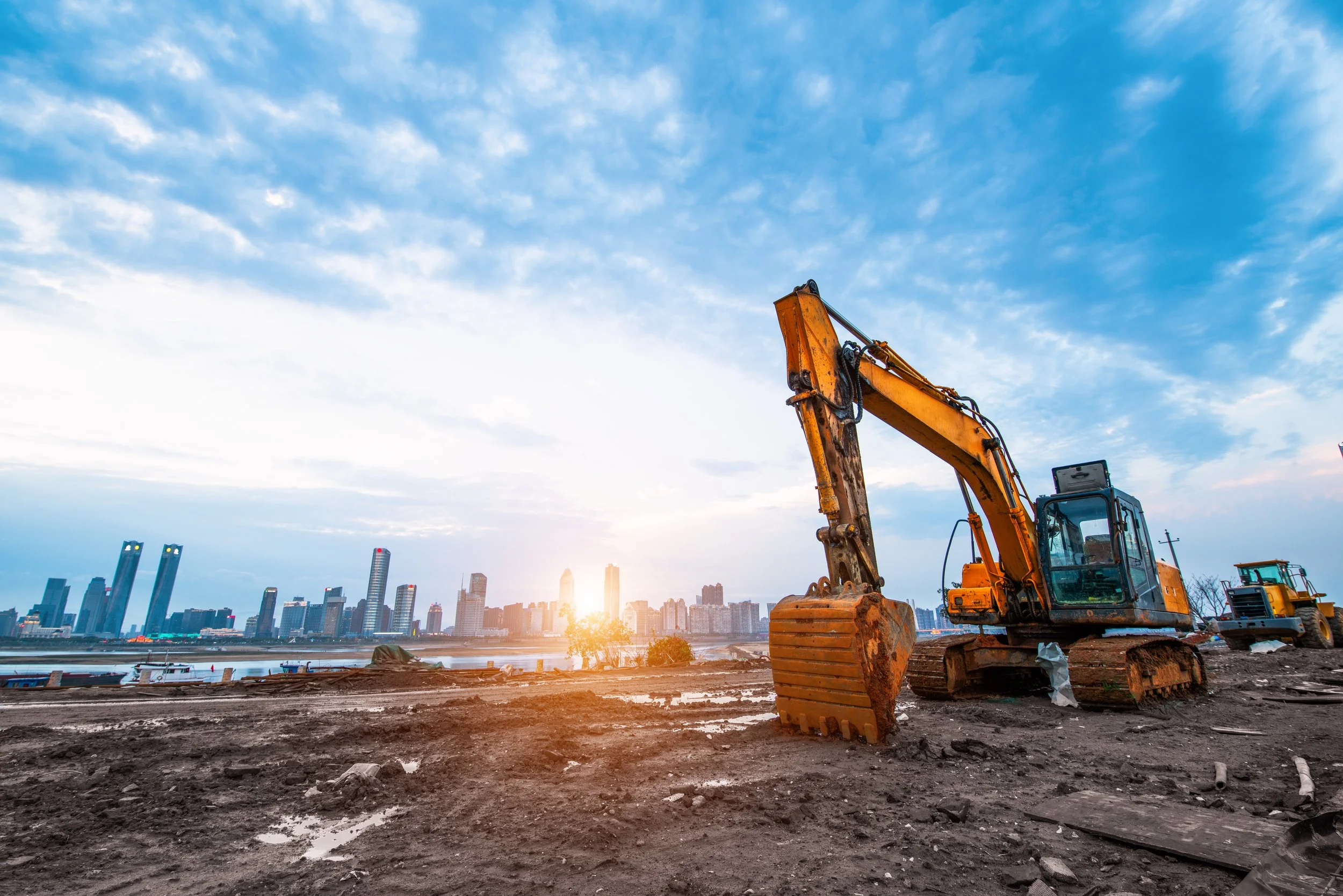 Construction site with a yellow excavator and a city skyline in the background under a partly cloudy sky during sunset.