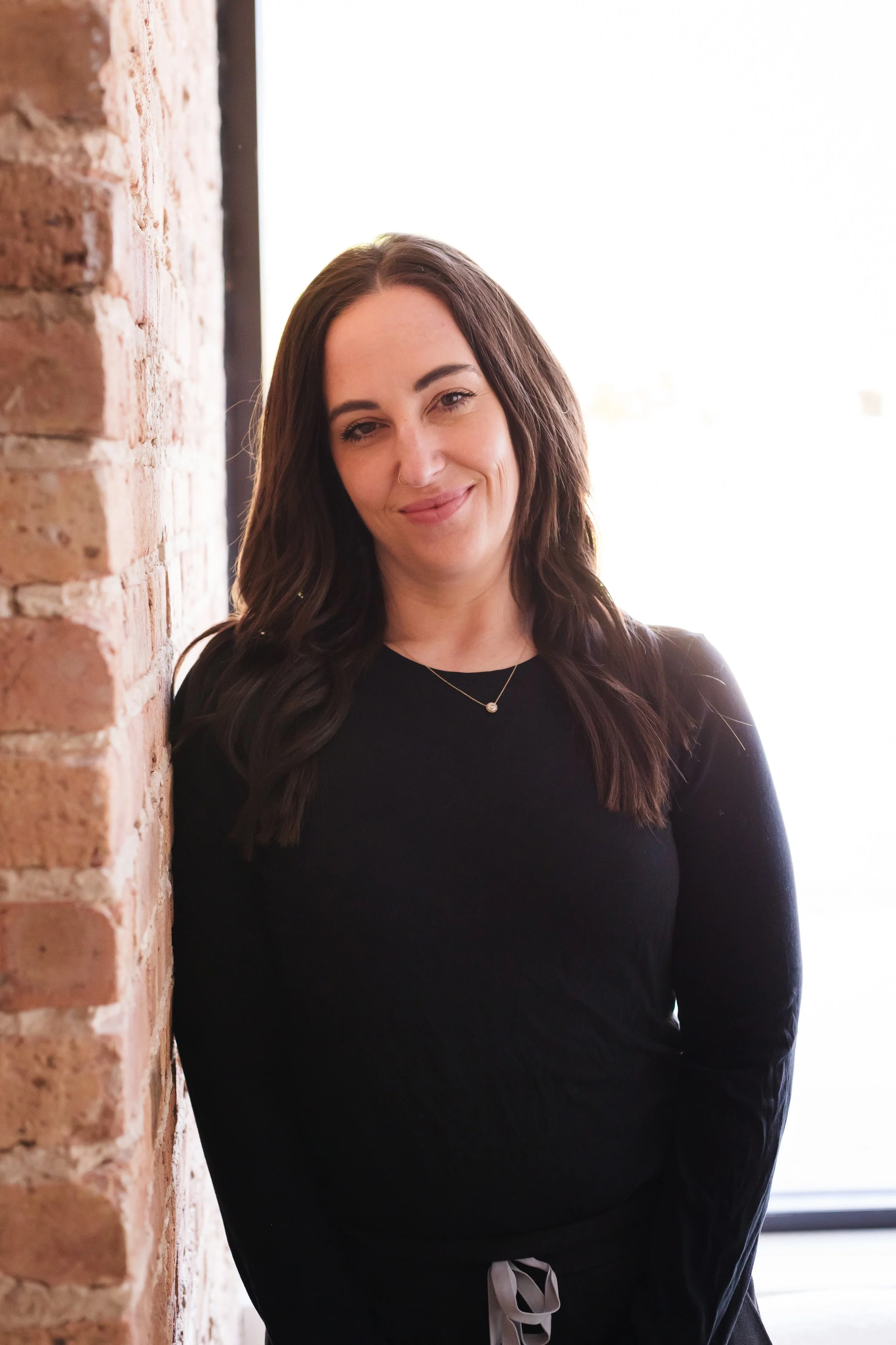 A woman with long dark hair wearing a black top and a necklace, standing next to an exposed brick wall, smiling at the camera with a bright background.