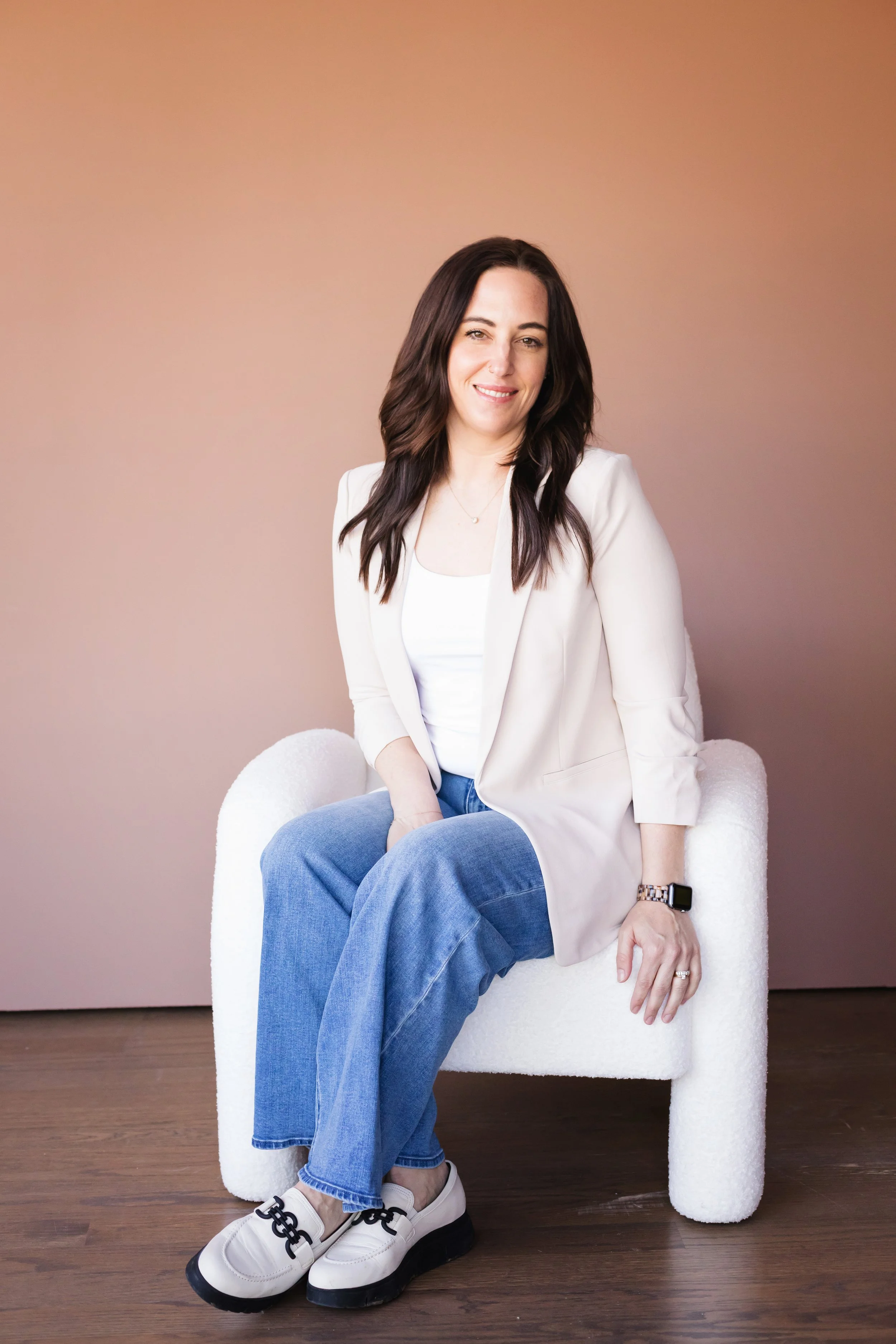 A woman with shoulder-length dark hair, wearing a light-colored blazer, white top, blue jeans, and white sneakers, sitting on a white plush chair against a beige background.