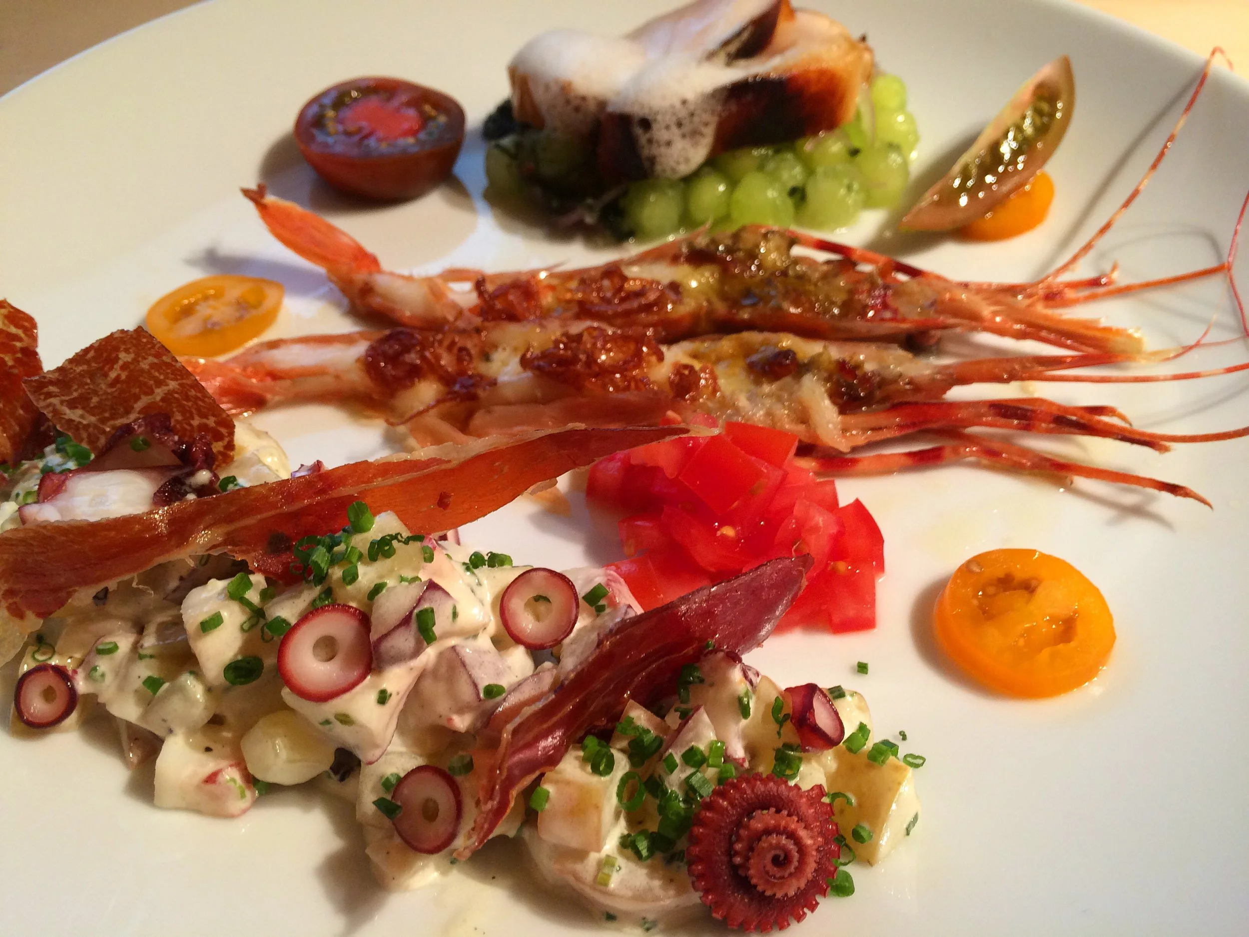 A white plate with various seafood and vegetable dishes, including lobster, sliced cherry tomatoes, radish slices, a halved cherry tomato, and a lobster tail, garnished with chopped herbs.