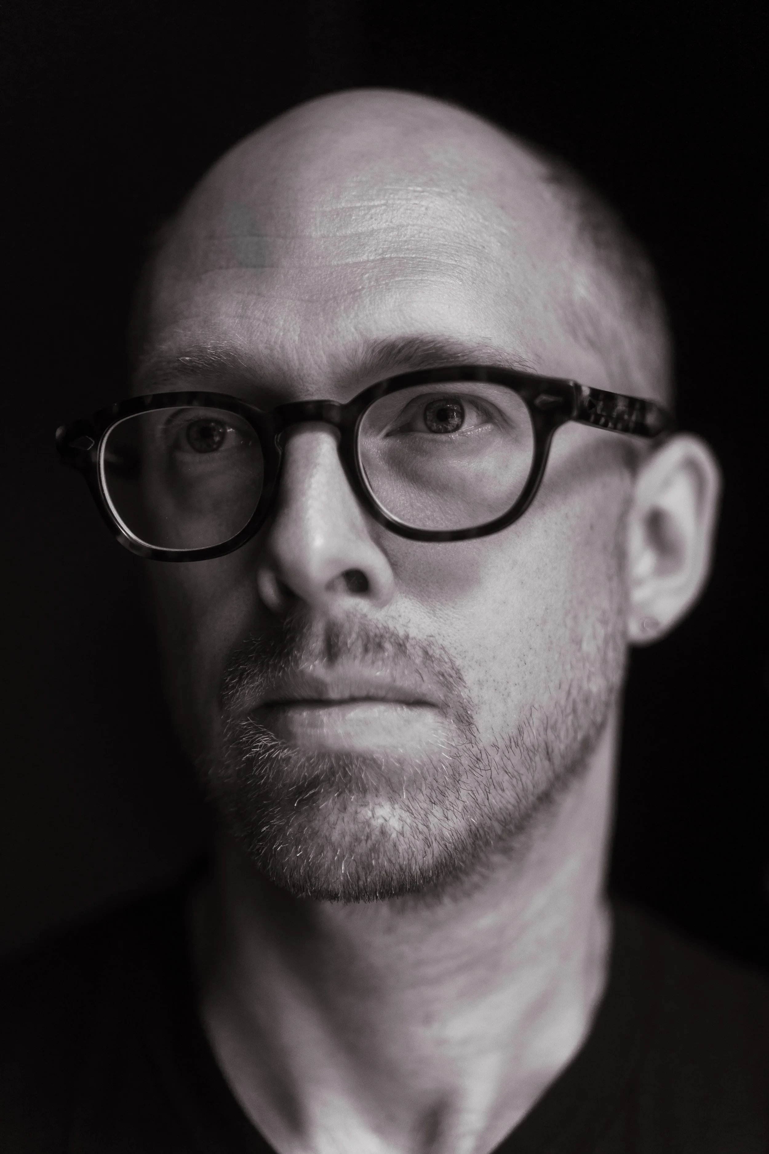 Black-and-white portrait of a man with a shaved head, glasses, and a beard, looking slightly downward with a serious expression.