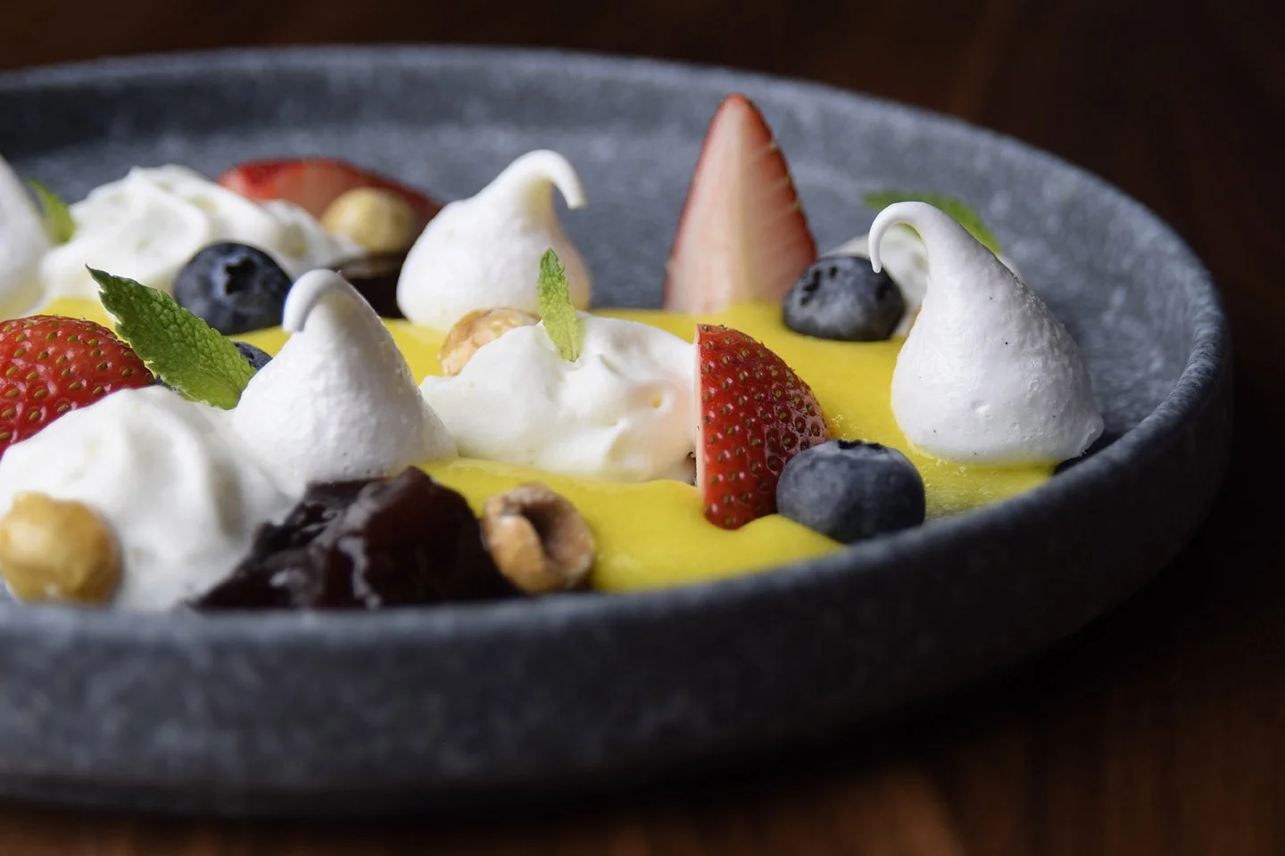 Close-up of a dessert with strawberries, blueberries, whipped cream, mint leaves, and a yellow sauce on a black plate.