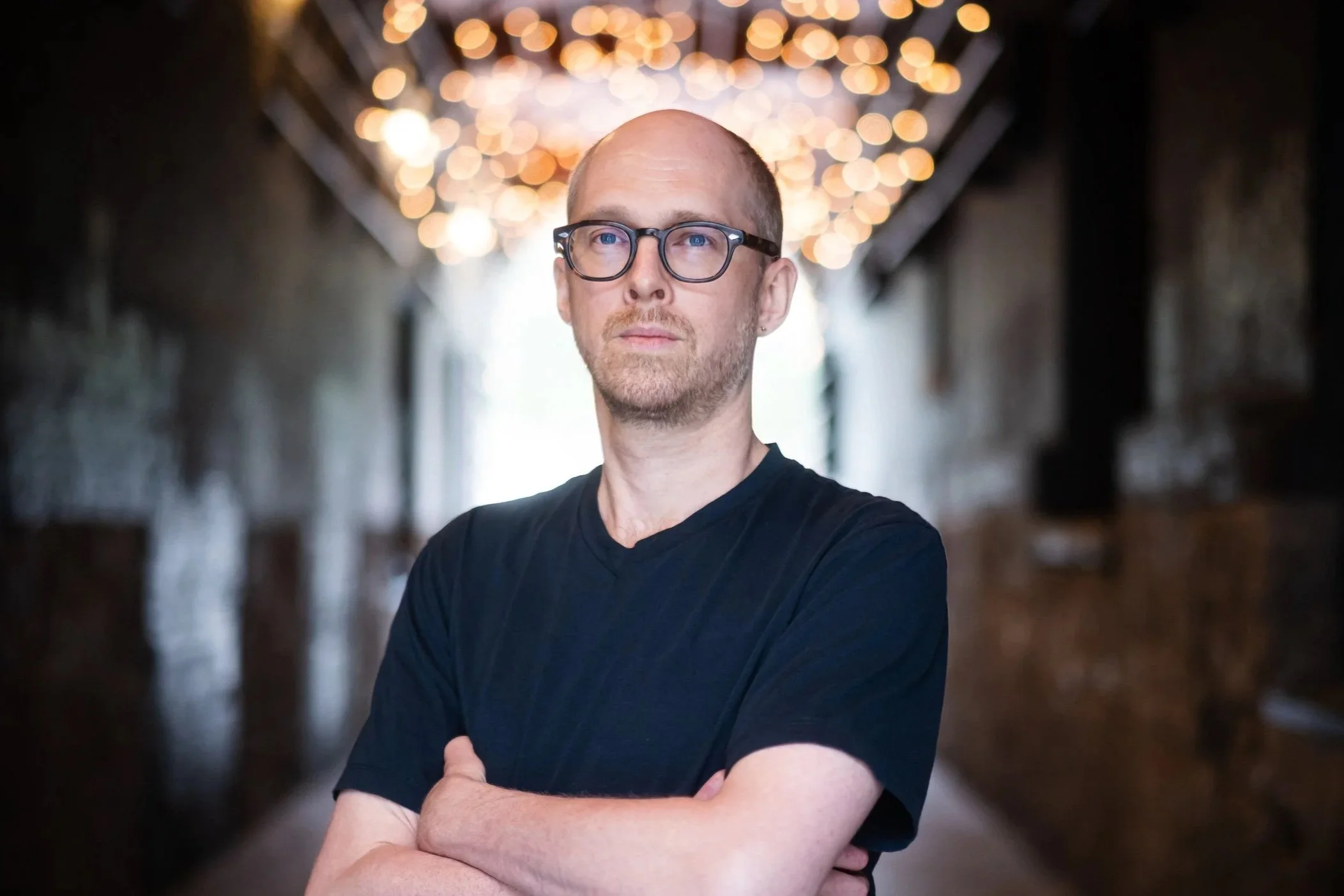A man with glasses and a shaved head standing with arms crossed in a corridor with warm, hanging string lights behind him.