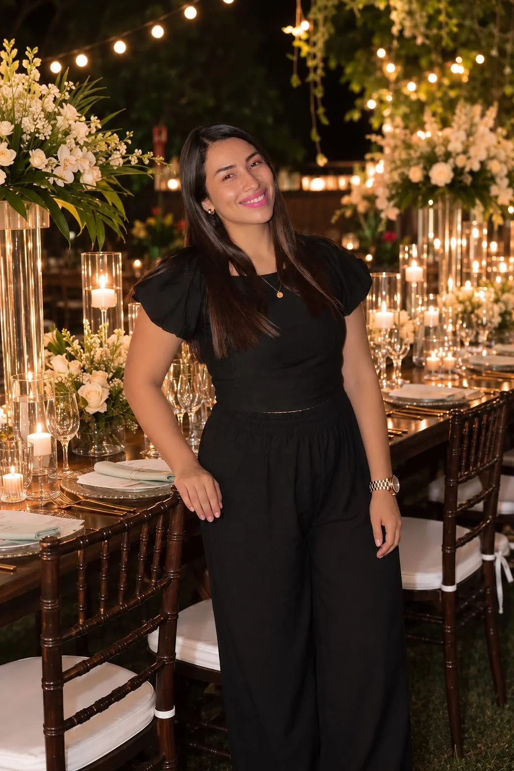 A woman in black outfit smiling at an elegant outdoor dinner celebration with floral arrangements and candlelit lighting.
