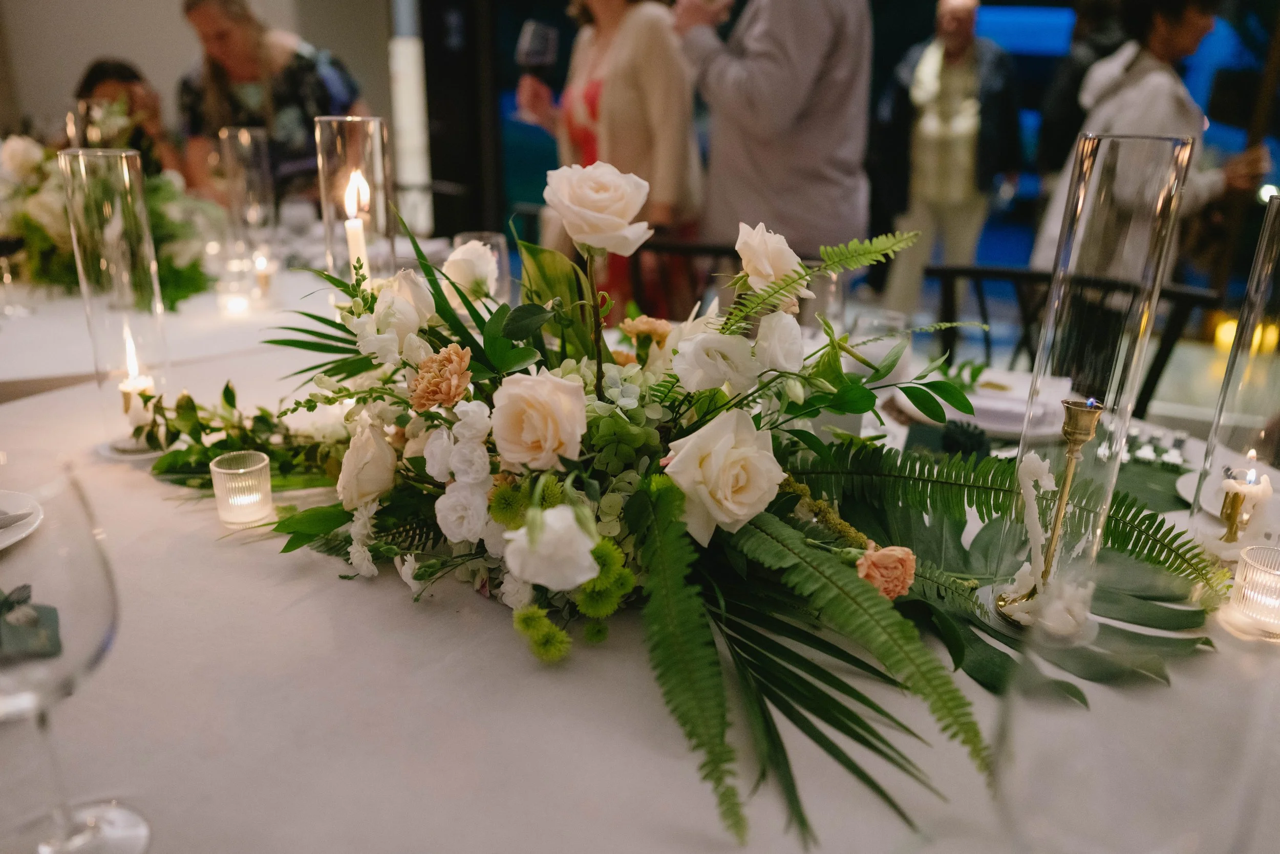 A floral centerpiece with white roses, peach flowers, and green ferns on a decorated table at an indoor event.