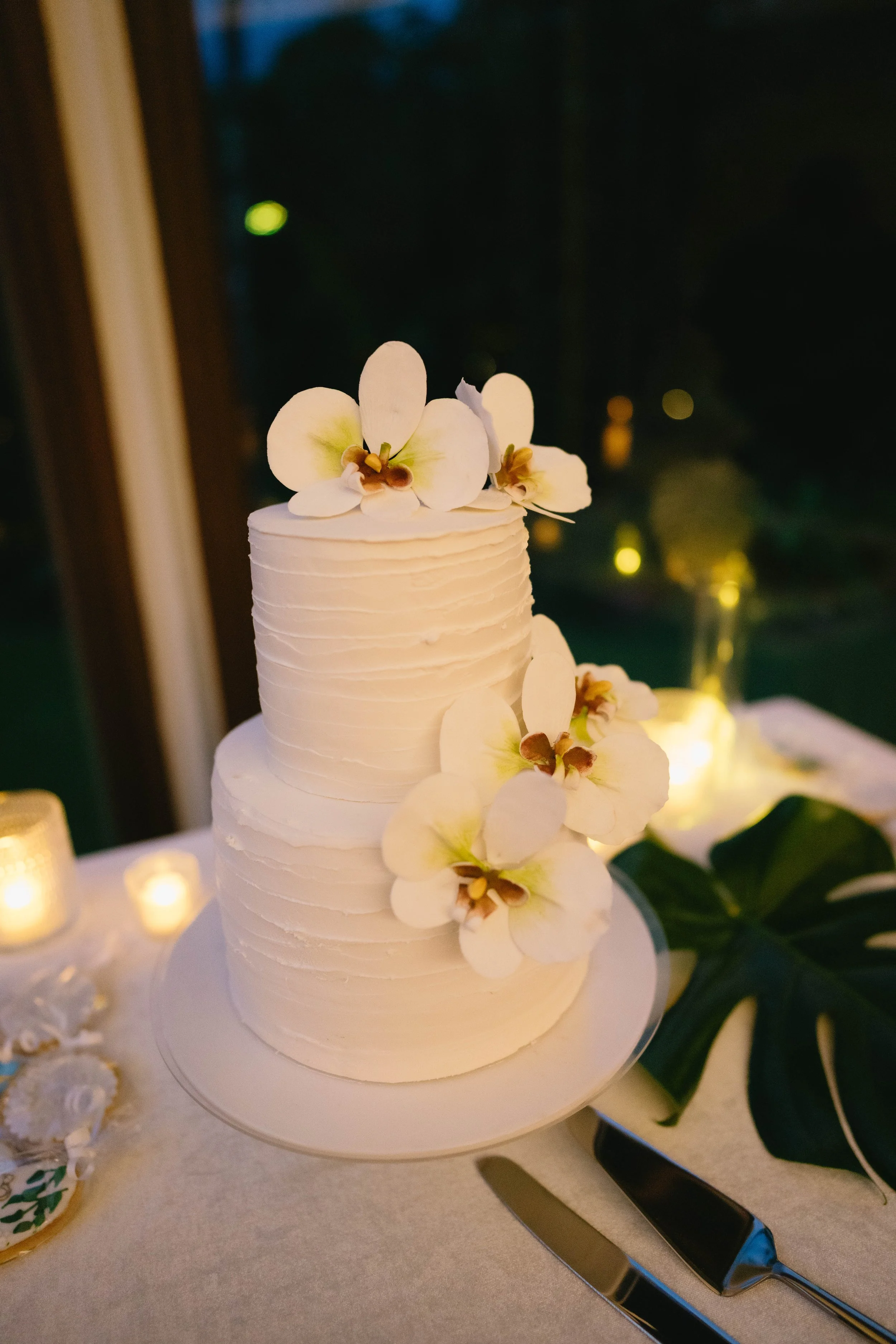 A white, three-tiered wedding cake decorated with cream-colored orchid flowers, placed on a white cake stand, with a table setting including a knife and cake server in front of it, and lit candles and greenery surrounding it.
