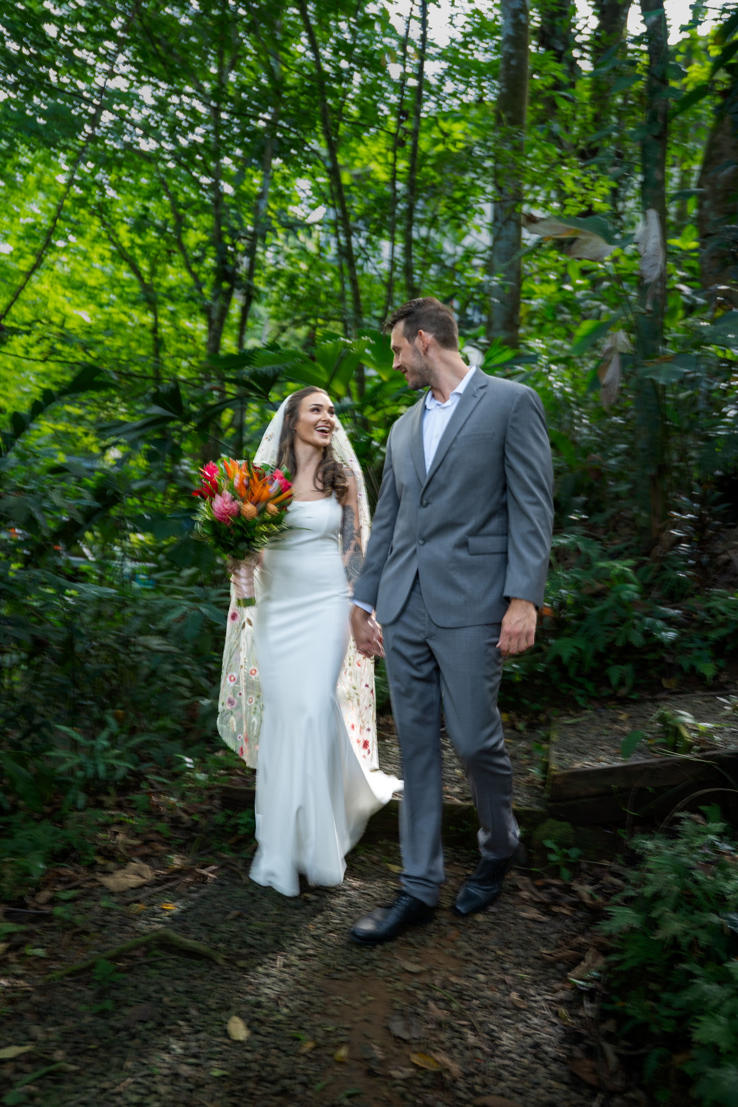 A bride and groom hold hands and walk through a lush green forest, smiling at each other on their wedding day. The bride wears a white wedding dress and veil, holding a bouquet of vibrant flowers, while the groom wears a gray suit.