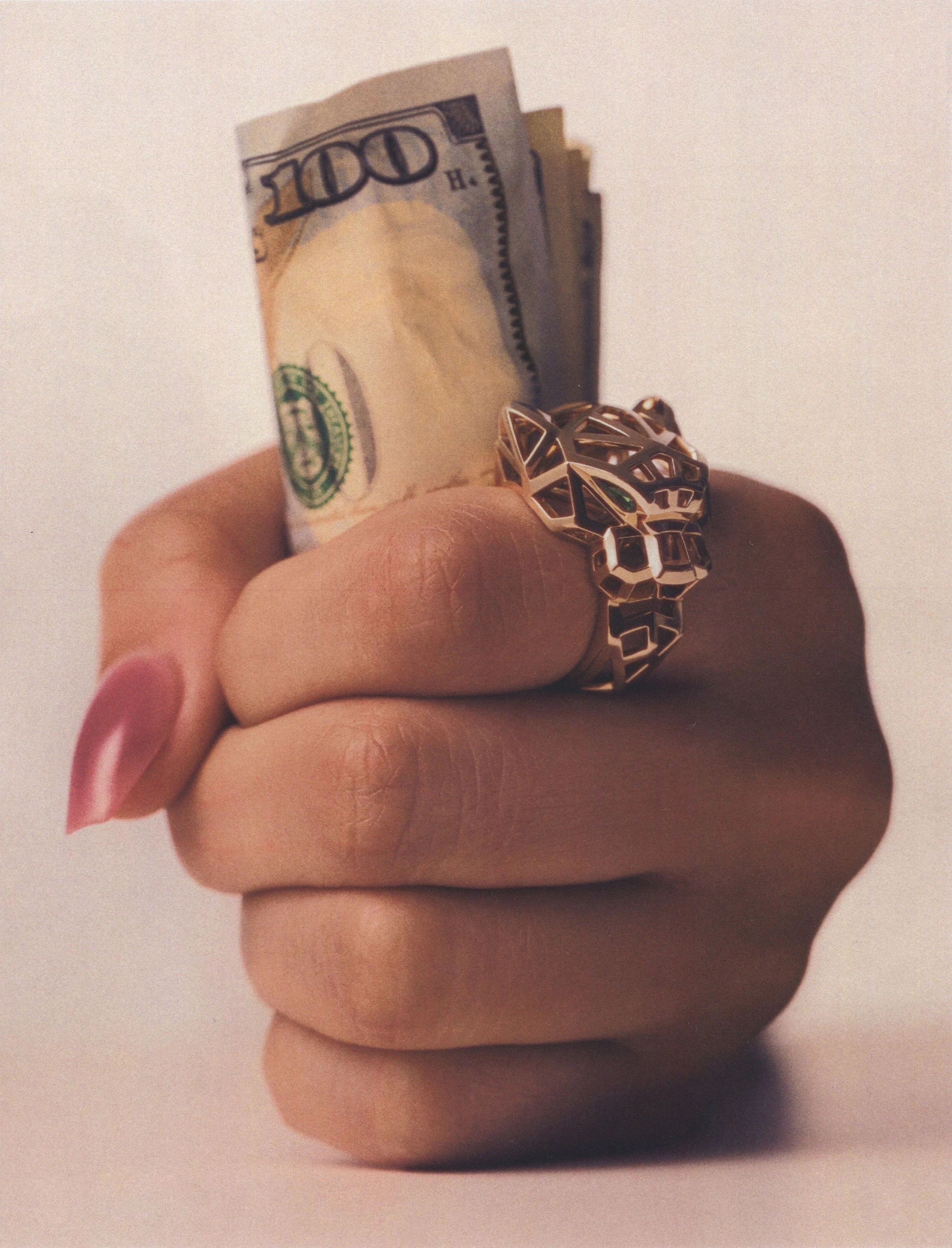 Close-up of a hand holding folded $100 bills while wearing a large gold panther ring.