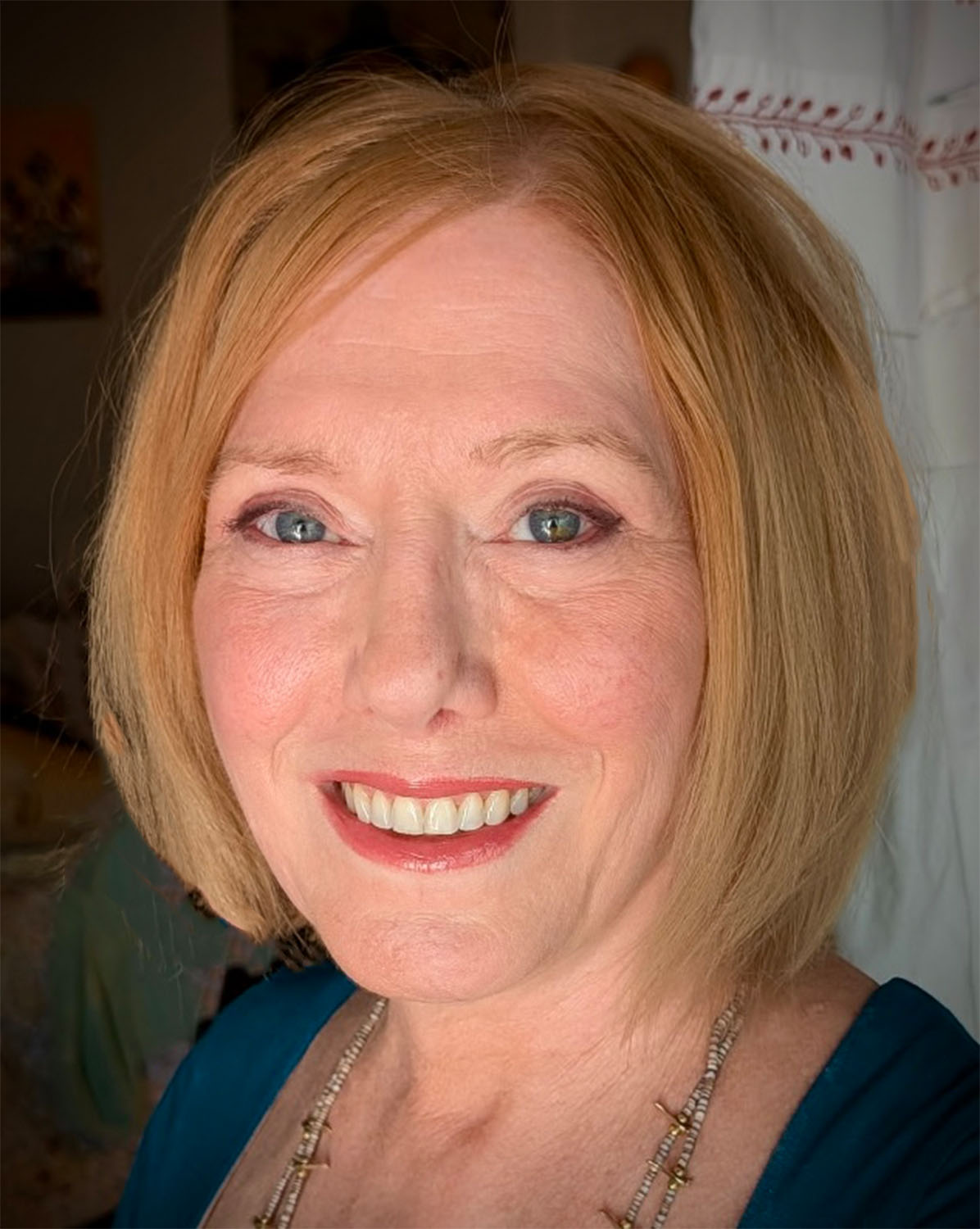A woman with blonde hair styled in a bob, smiling showing her teeth, wearing a blue top, a necklace, and earrings.