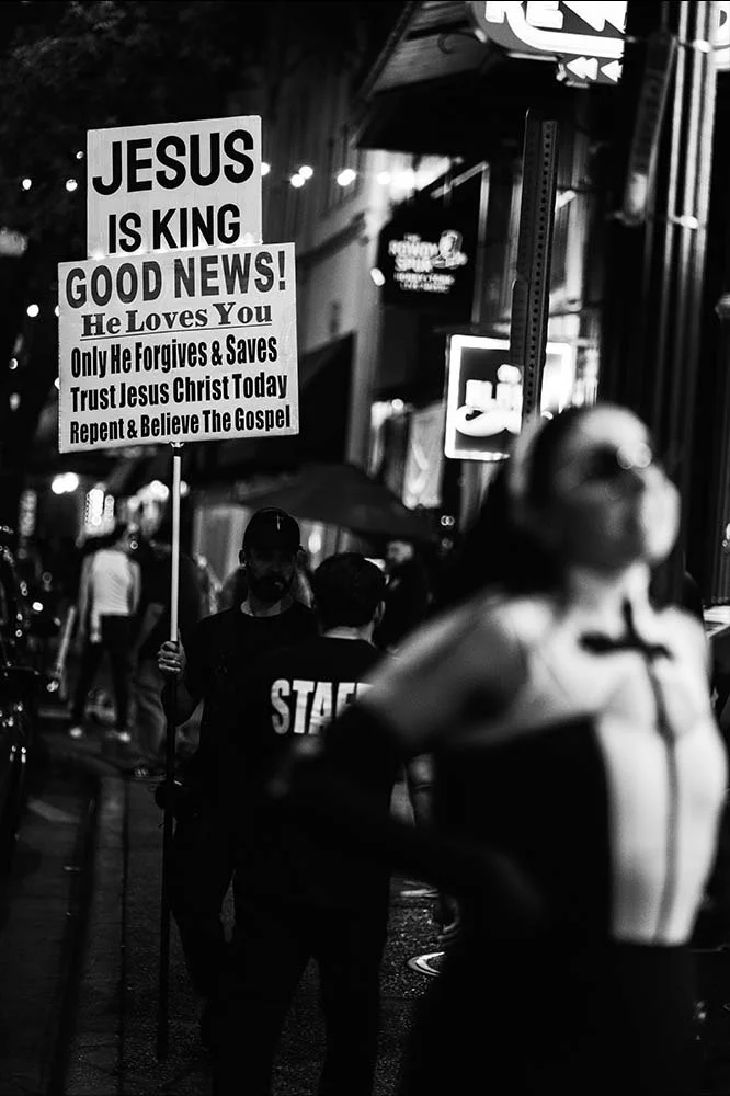 A person holding a sign that reads "Jesus is King Good News! He Loves You" on a busy city street at night.