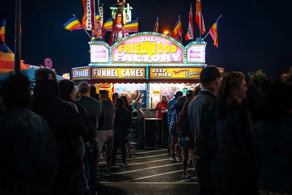 Nighttime scene at a fair with a food stand called 'The Fried Food Factory' and a line of people waiting. The stand is decorated with colorful flags and bright lights, offering funnel cakes, deep-fried Twinkies, and Oreos.