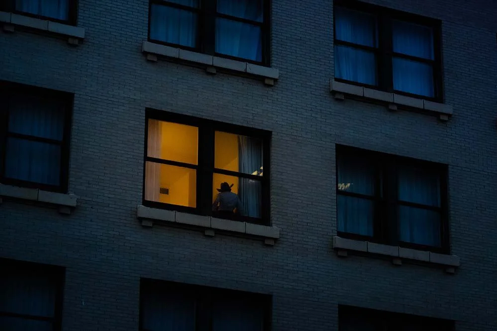 A person wearing a cowboy hat sitting in a warmly lit window of an apartment building at dusk.