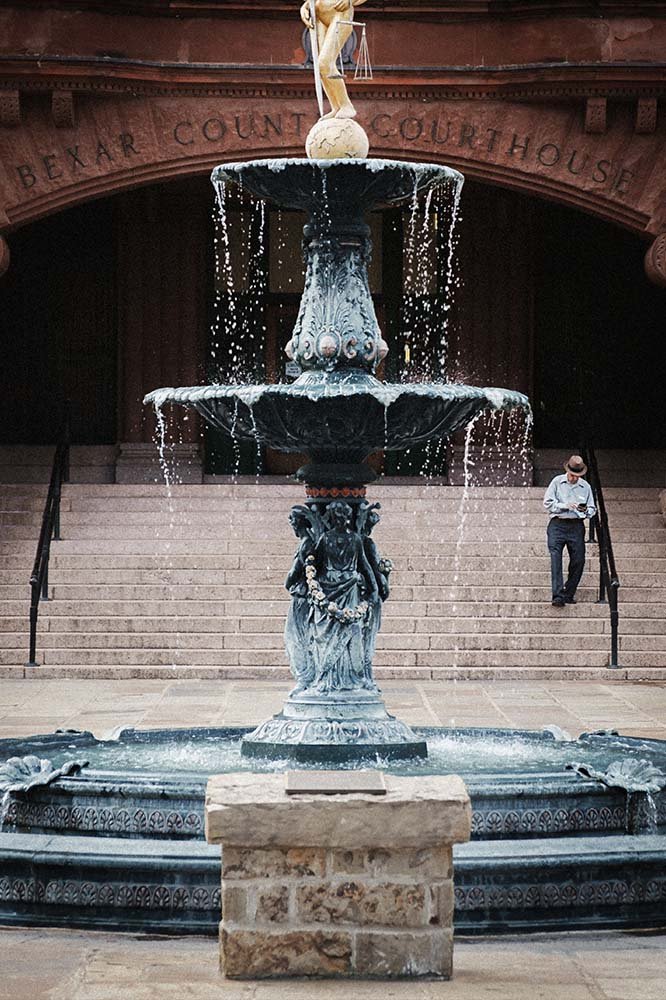A historic fountain with sculptures in front of the Bexar County Courthouse, featuring water flowing from multiple tiers, with steps and a person standing on the stairs.