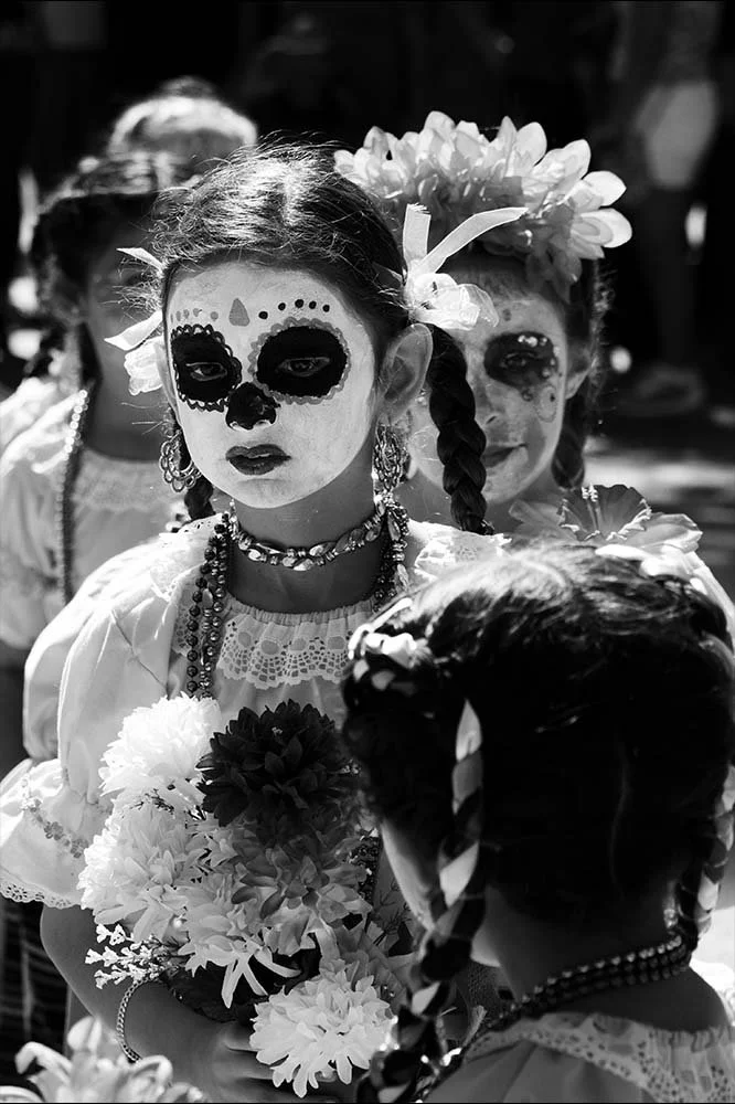 Girls with Día de los Muertos face paint, wearing traditional dresses, and braided hair decorated with flowers, participating in a celebration.