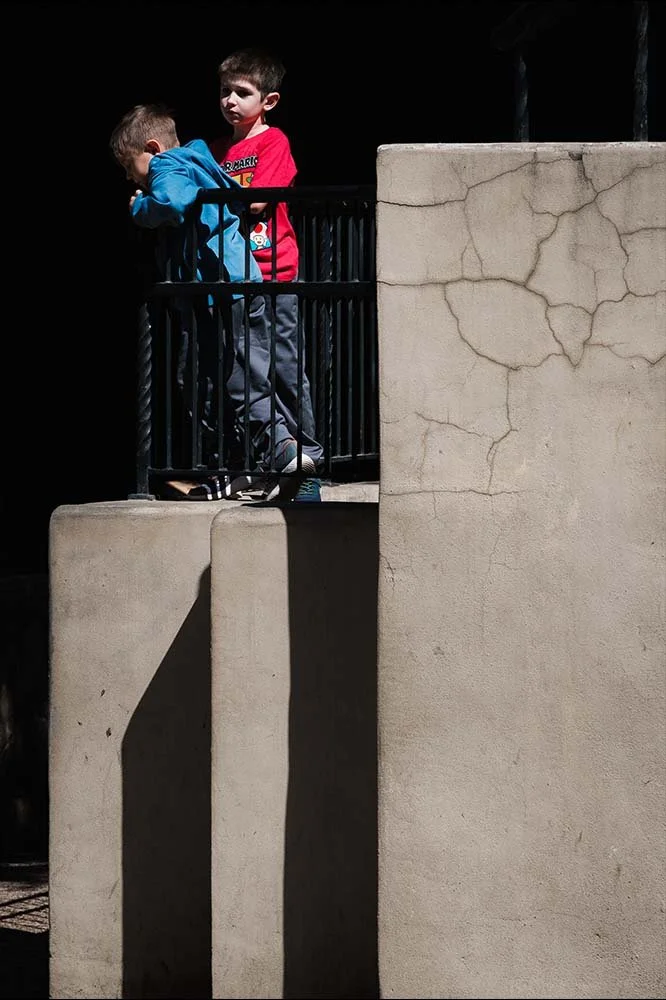 Two young boys standing on a balcony with black railings, one wearing a blue hoodie and the other in a red shirt, against a background of a black wall and a cracked beige wall.