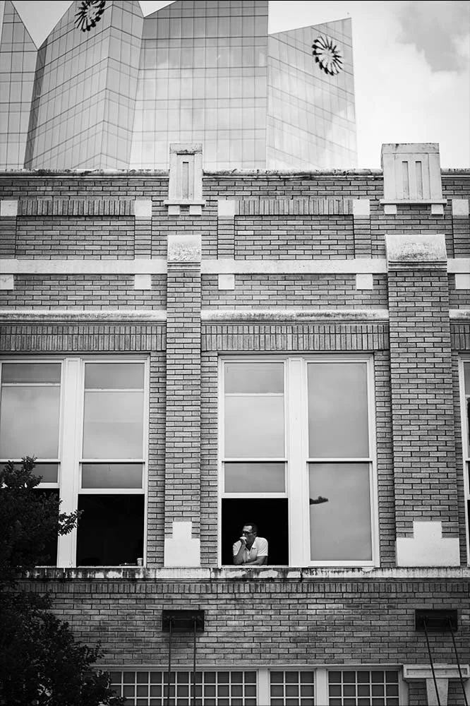 A person standing by a large window looking at a cellphone, in a brick building with multiple large windows, with a modern glass skyscraper with reflective surfaces and flower-shaped patterns on its upper part in the background.