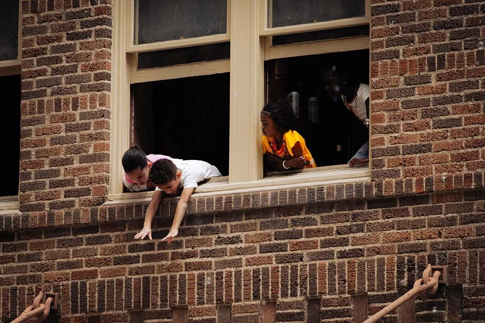 Two children leaning out of a brick building window, with a woman looking from inside.