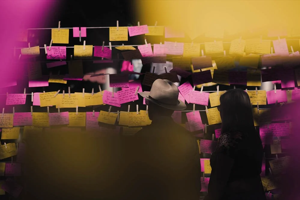 Two people viewing a wall of colorful notes or messages attached with clothespins.