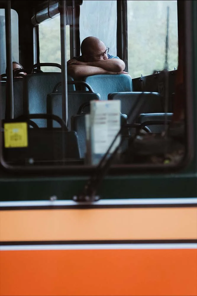 A person with glasses sleeping on a bus, resting their head on their arm, looking out the window.