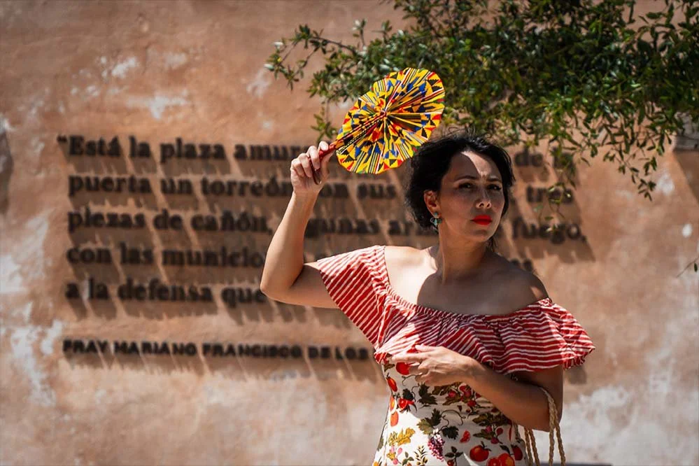 A woman holding a colorful decorative fan in front of a wall with a Spanish quote.