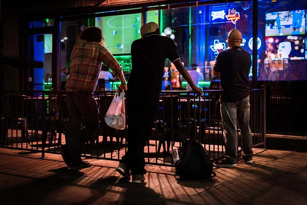 Three men are watching a large sports game on multiple screens at an entertainment bar or sports pub during nighttime. They are standing behind a black metal fence, with one holding a plastic bag and another with a backpack on the ground.