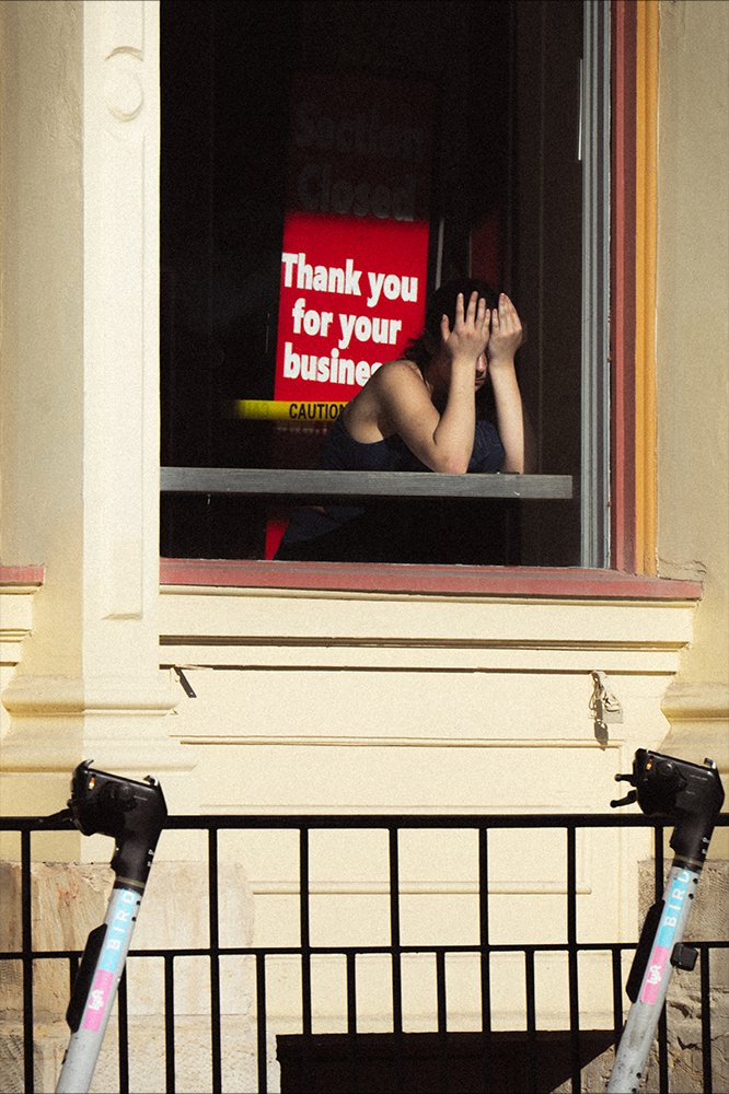 A woman sitting inside a building behind a window, covering her face with her hands, with a sign behind her that reads 'Thank you for your business.' Two electric scooters are parked in front of the window.