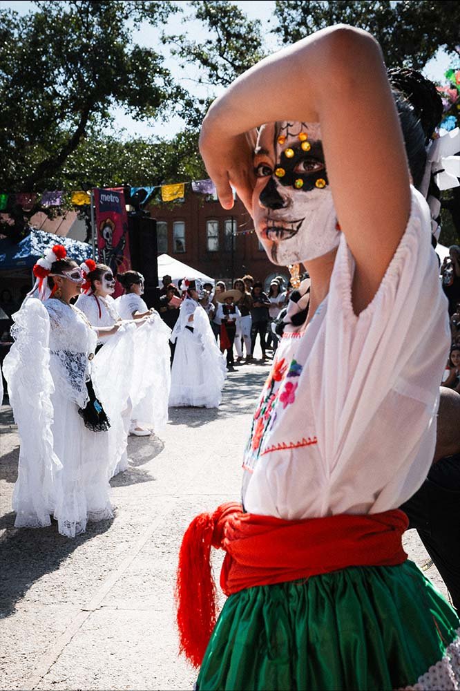 Child dressed in traditional Mexican clothing with face painted as a calavera, participating in a cultural celebration or parade, with other children in white dresses and decorated face paint in the background.