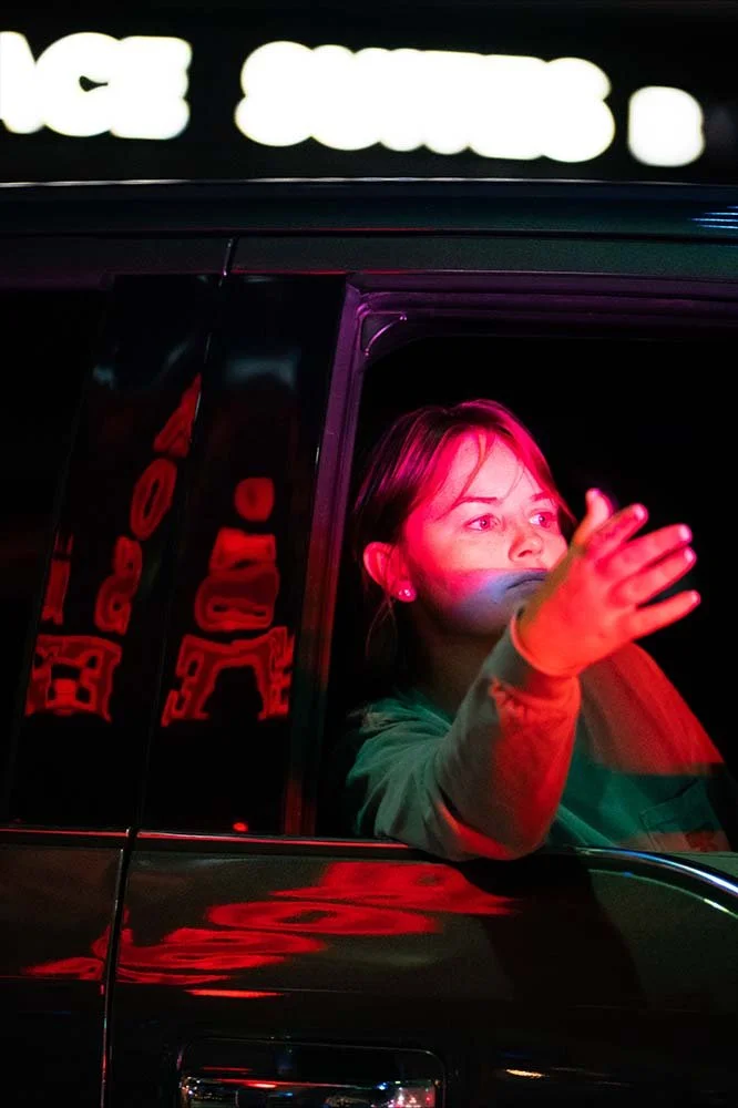 Woman with dark hair waving from car window at night, illuminated by red and pink neon lights.