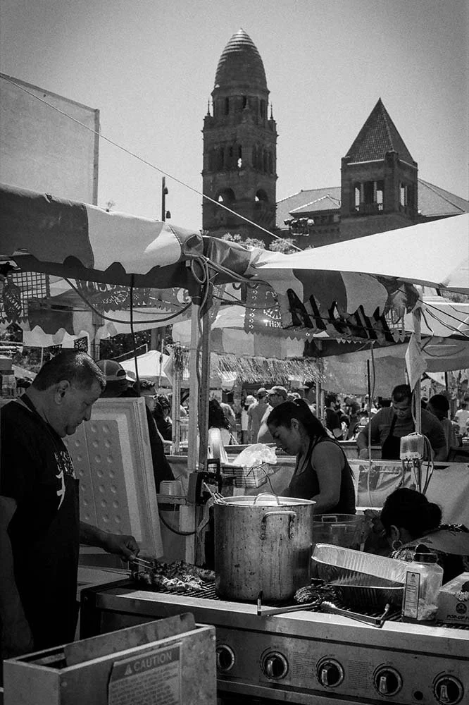 Black and white photo of an outdoor market with tents, vendors, and people. A large pot and grill with skewers of meat are in the foreground. In the background, there is a tall, historic church tower with a spire and a steep-roofed building.