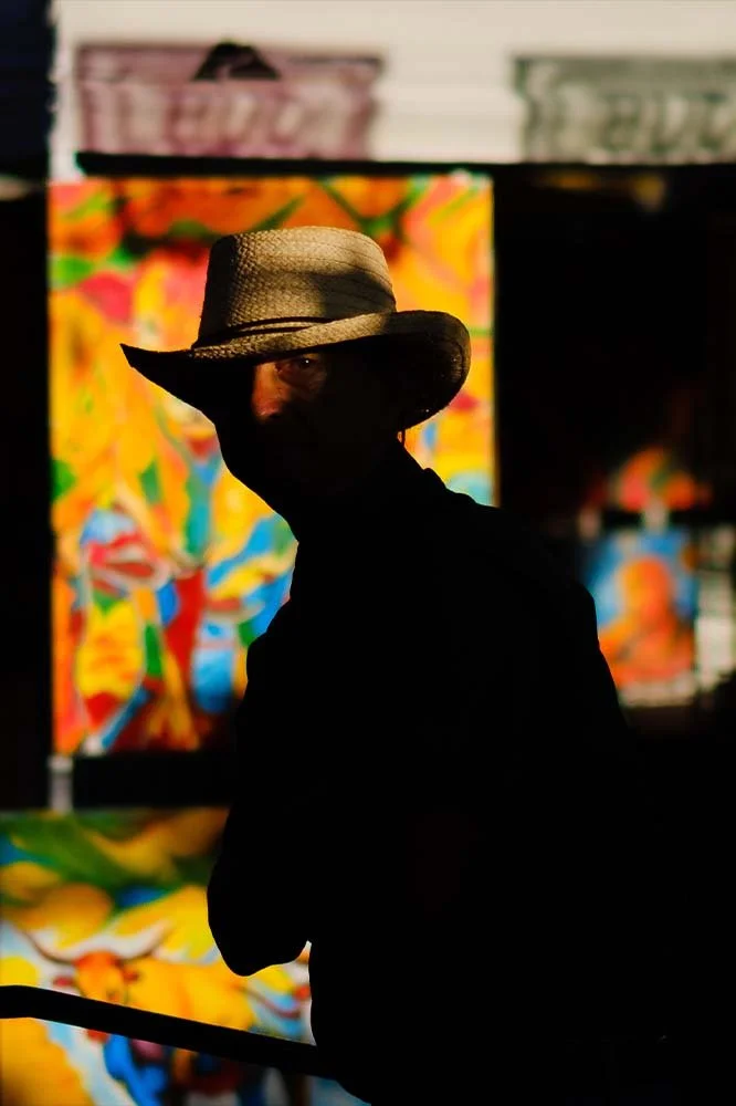 Person wearing a wide-brimmed straw hat and face mask, in front of colorful abstract art or mural.
