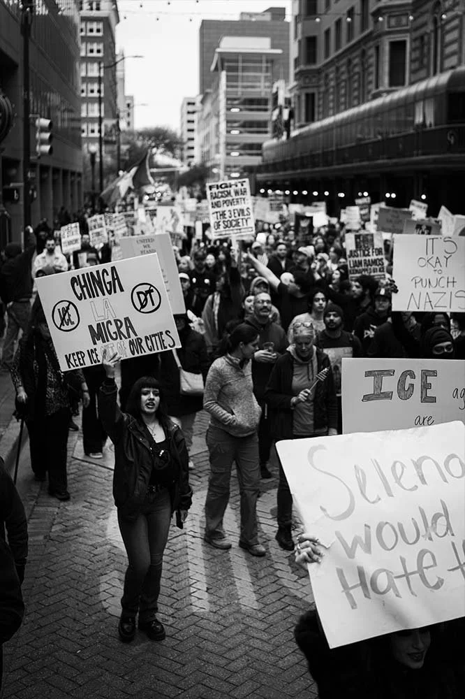 Crowd of people participating in a protest march on city street, holding various signs advocating against racism, misogyny, and hate crimes, with tall buildings and city infrastructure in the background.