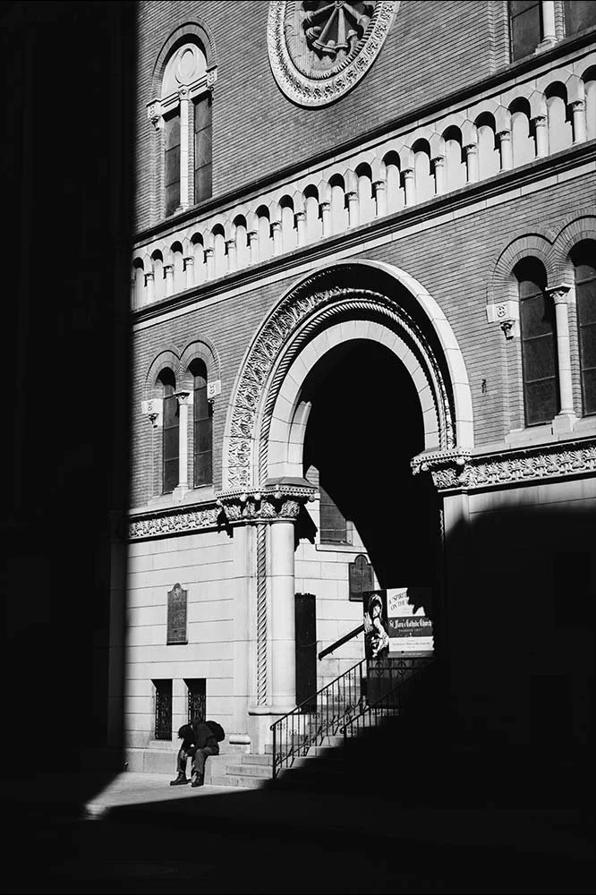 Black and white photo of a historic brick building with an arched entrance, decorative columns, and a clock on the upper facade. Two children sit on the steps beneath the arch, with one climbing the stairs and a person walking near the entrance.