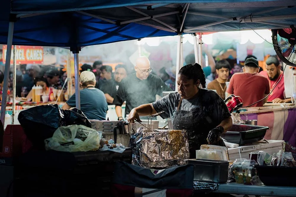 A street food stall at a busy fair or market with a woman cooking and serving food, surrounded by crowds of people and colorful stalls.