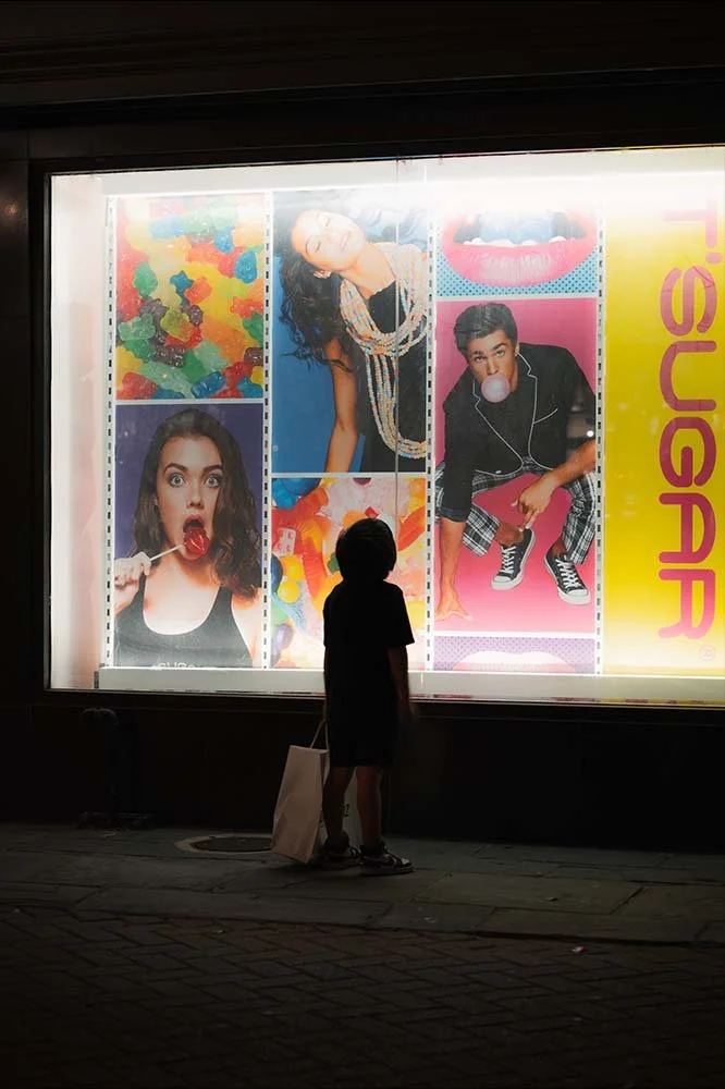 A young boy standing outside at night, looking at a brightly lit store window display with colorful images of people eating candy, blowing bubbles, and posing for photos.