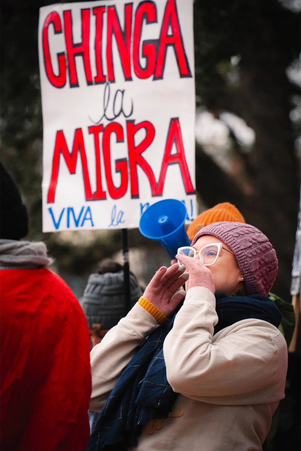 A woman wearing a pink beanie, glasses, and a beige jacket is speaking into a megaphone at a protest, with a sign in the background that reads 'Chinga la MIGA' and 'Viva la' partially visible.