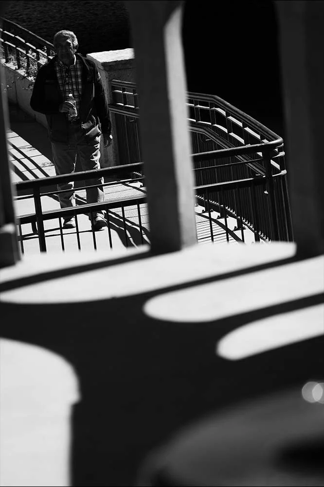 A man walking on a spiral staircase holding a water bottle, seen through black railings and shadows, black and white photo.