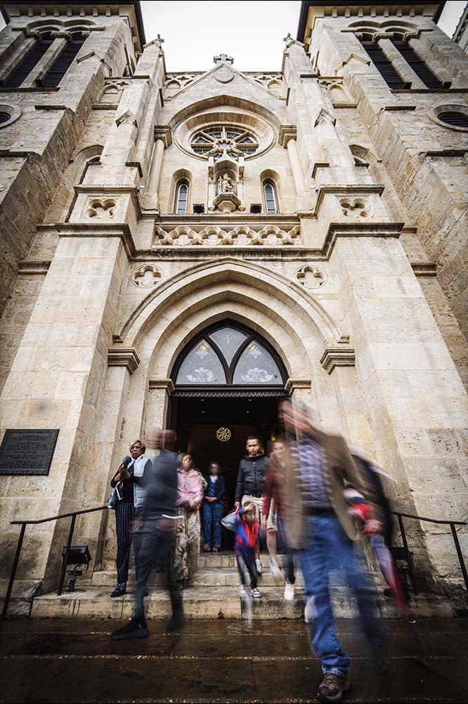 People exiting a historic church with gothic architecture, featuring arched entrance, stone facade, and large stained glass window above the door.