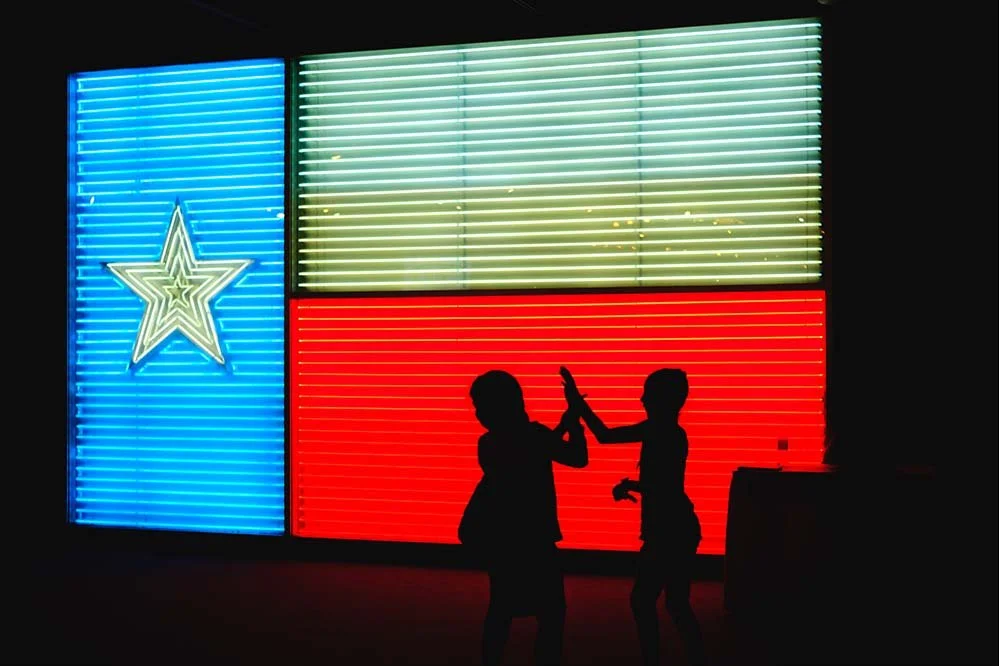 Silhouettes of two children playing in front of a neon Texas flag with a star, illuminated in blue, white, and red lights.