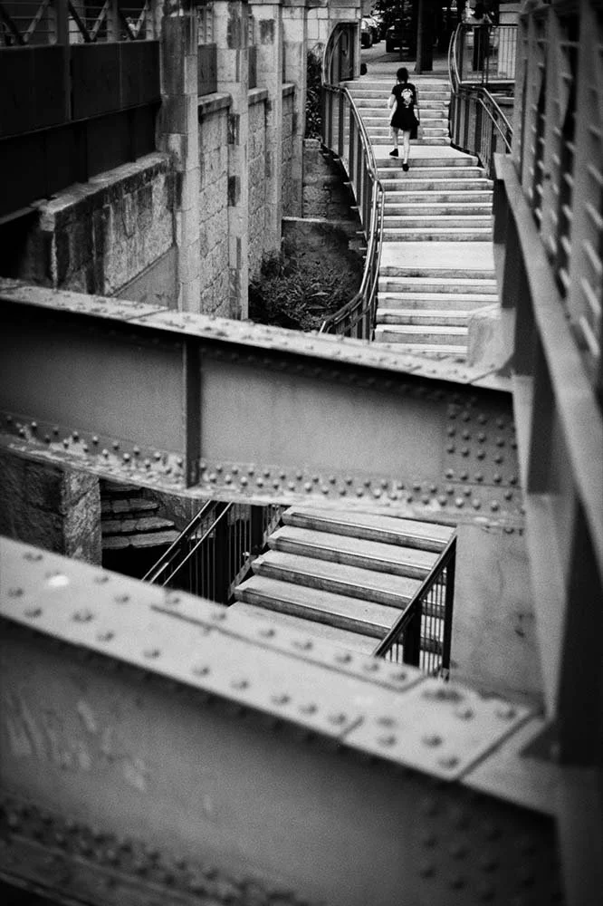 Black and white photo of a young girl walking upstairs on an outdoor staircase between buildings, shot through a metallic bridge or structure in the foreground.