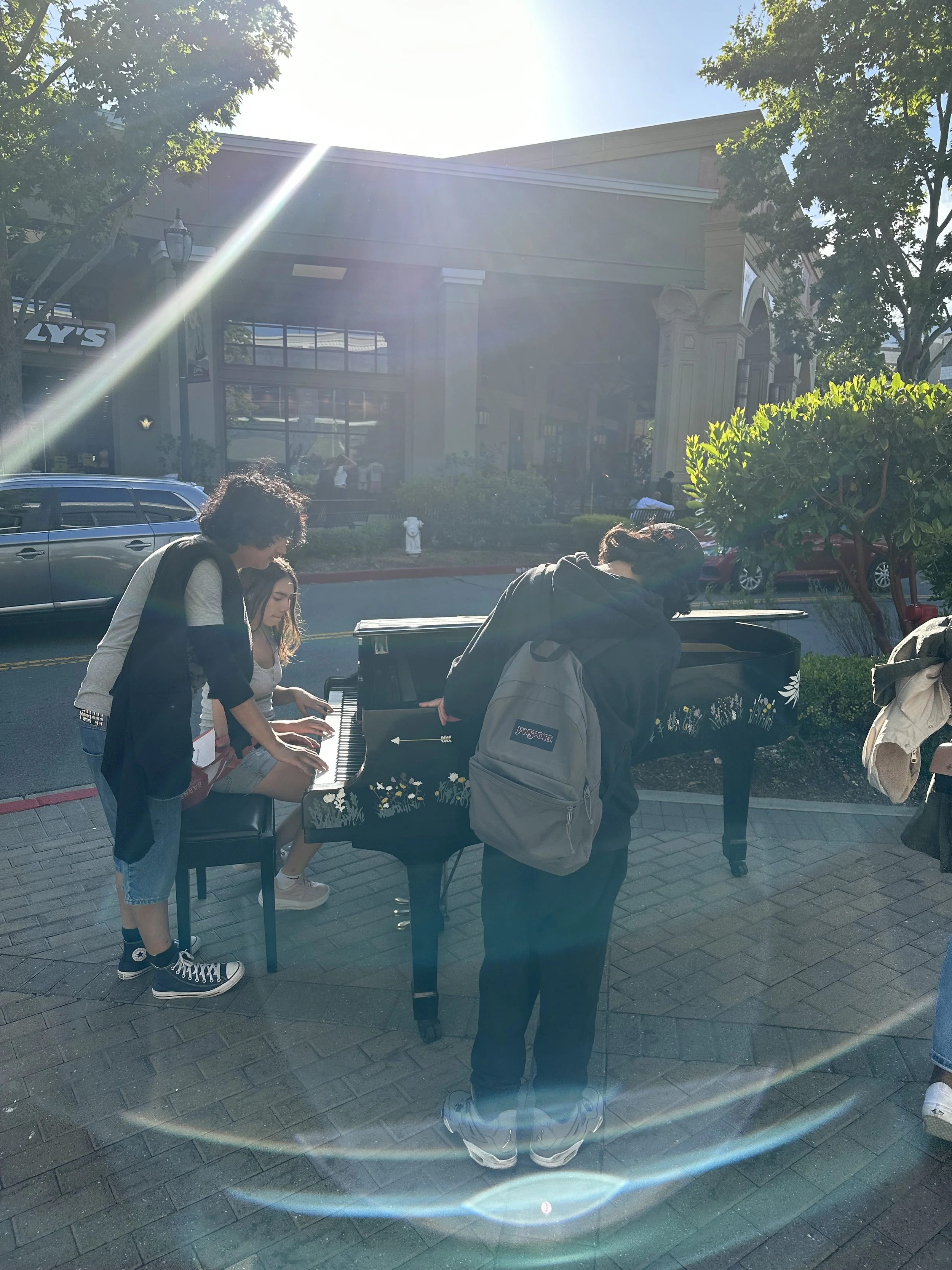 People playing piano outdoors in a shopping center parking lot on a sunny day, with trees and buildings in the background.