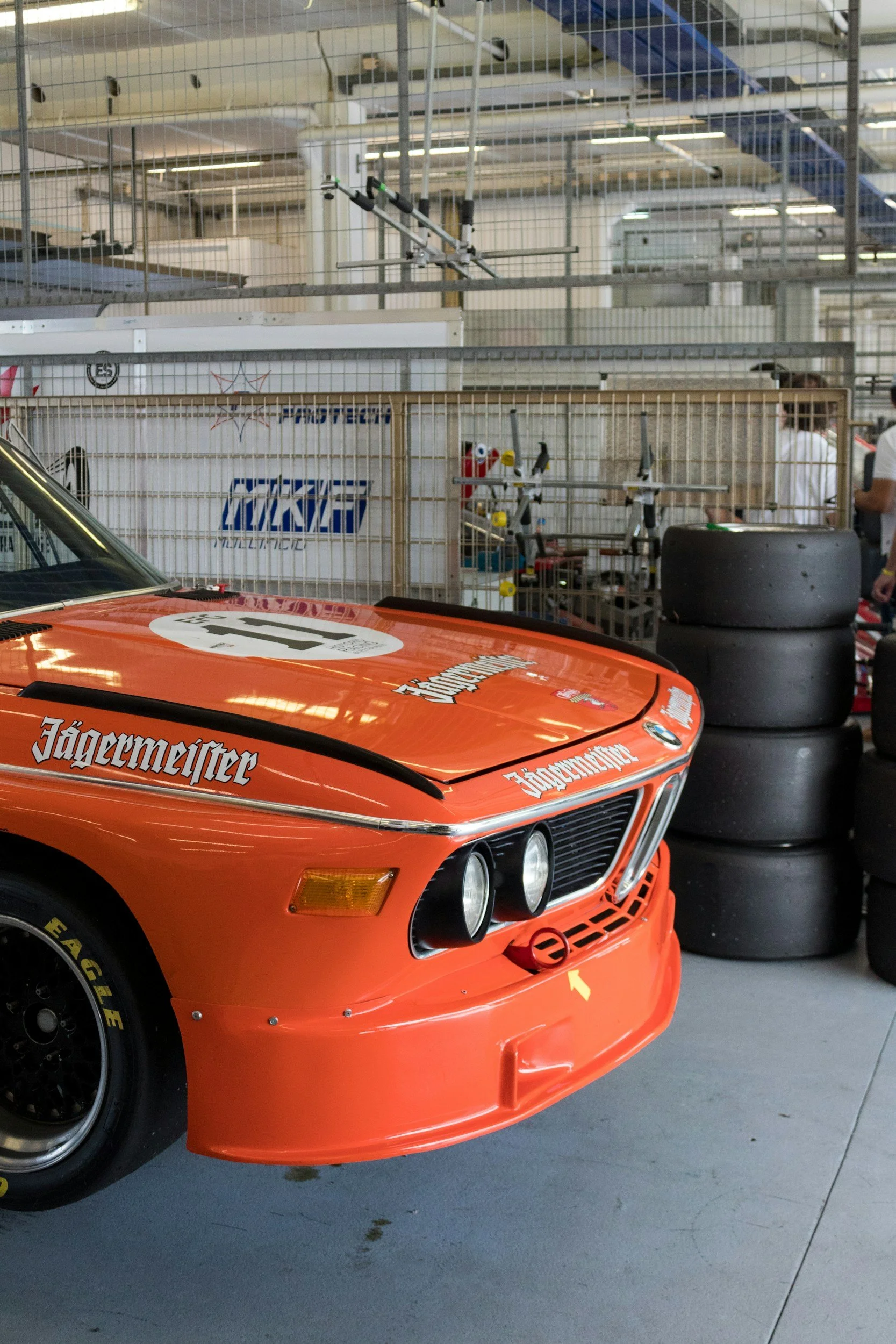 Close-up of an orange race car with Jägermeister branding, showing front grille, headlights, and tire, with stacked tires and fence in the background.