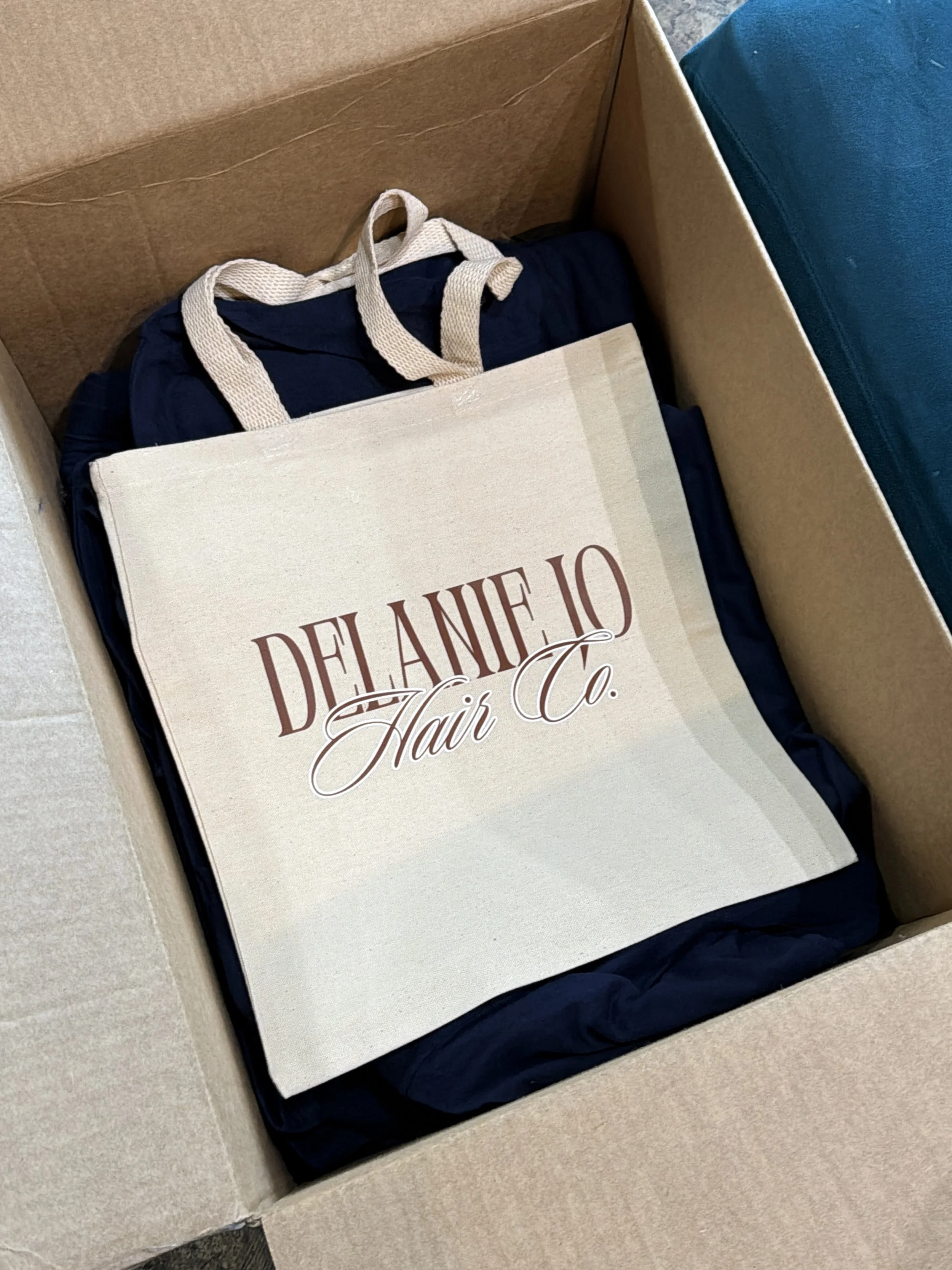 A cardboard box containing a navy blue tote bag with beige handles, a beige book titled "Delane O Hair Co." with decorative lettering, and a blue object, possibly a cushion or fabric. The items are arranged inside the box, which is lined with navy bl