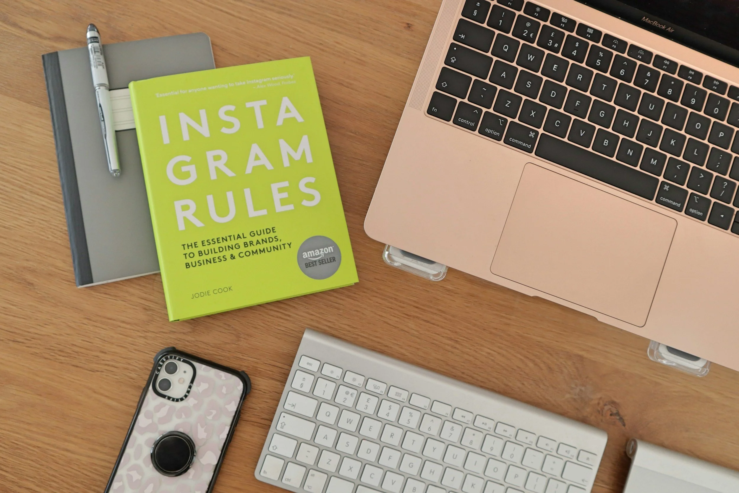 A wooden desk with a silver laptop and a white keyboard, an iPhone with a camouflage case, a yellow book titled 'Instagram Rules,' a gray notebook, and a pen.