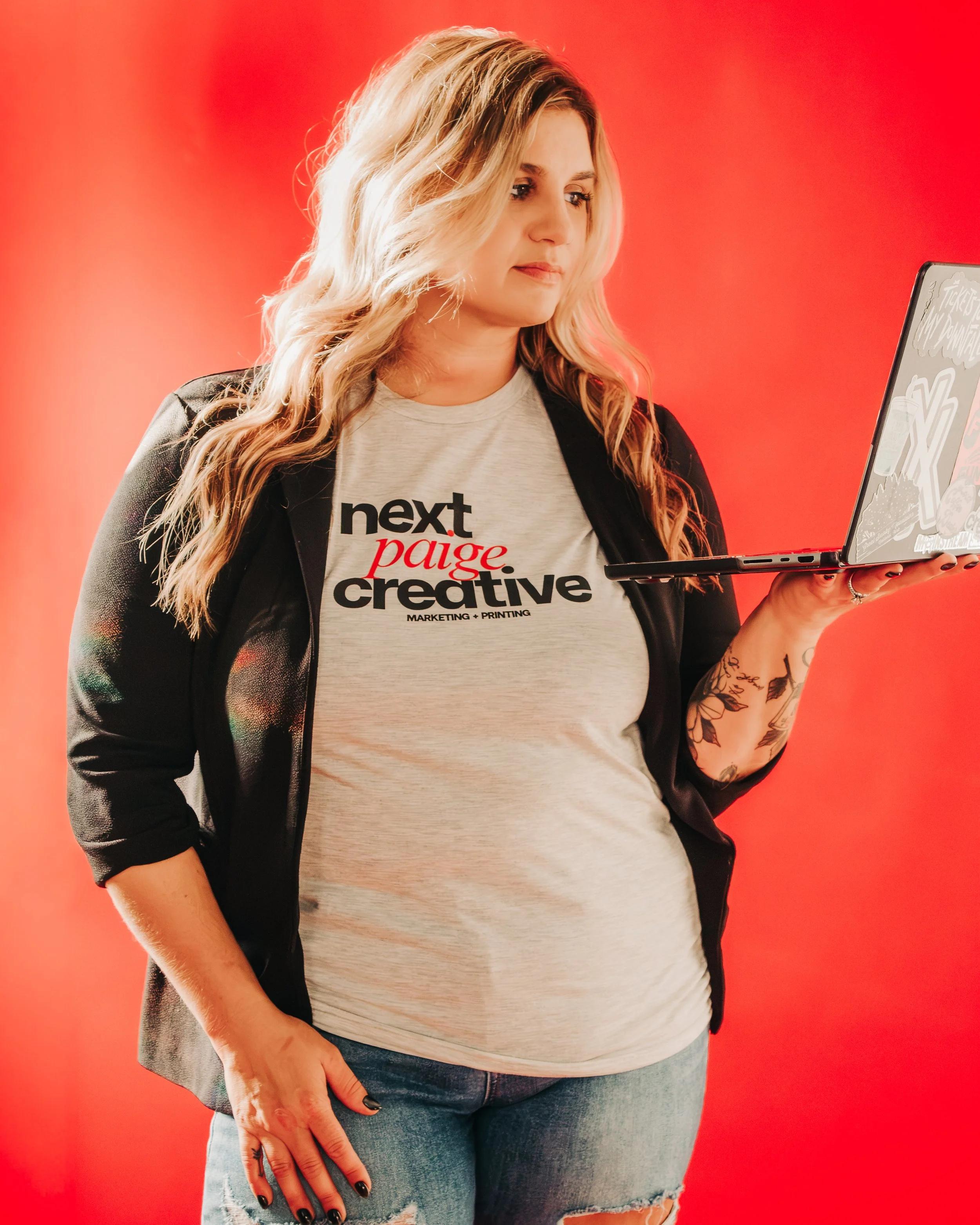 A woman with blonde wavy hair wearing a gray t-shirt with black and red text, a black jacket, and ripped jeans, standing against a red background while looking at her laptop.