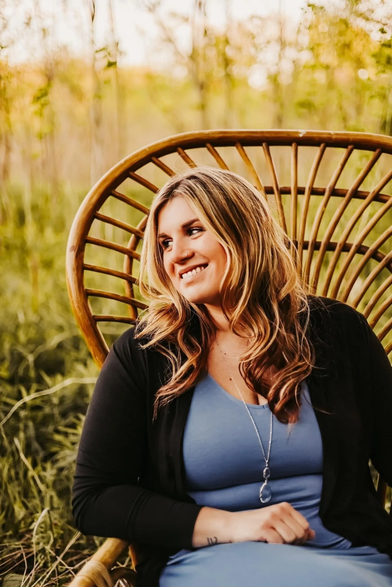 A young woman with long, wavy hair sitting outdoors on a wicker chair, surrounded by green foliage, smiling and looking to her right.