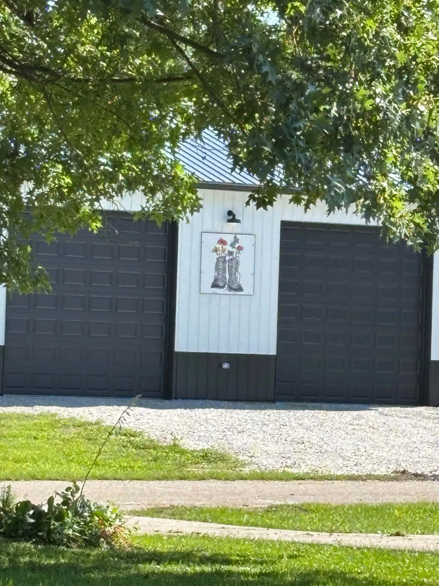 A white wall with two black garage doors, a painting of boots with flowers above an outdoor light fixture, green tree branches overhead, a gravel driveway, and a grassy lawn in the foreground.