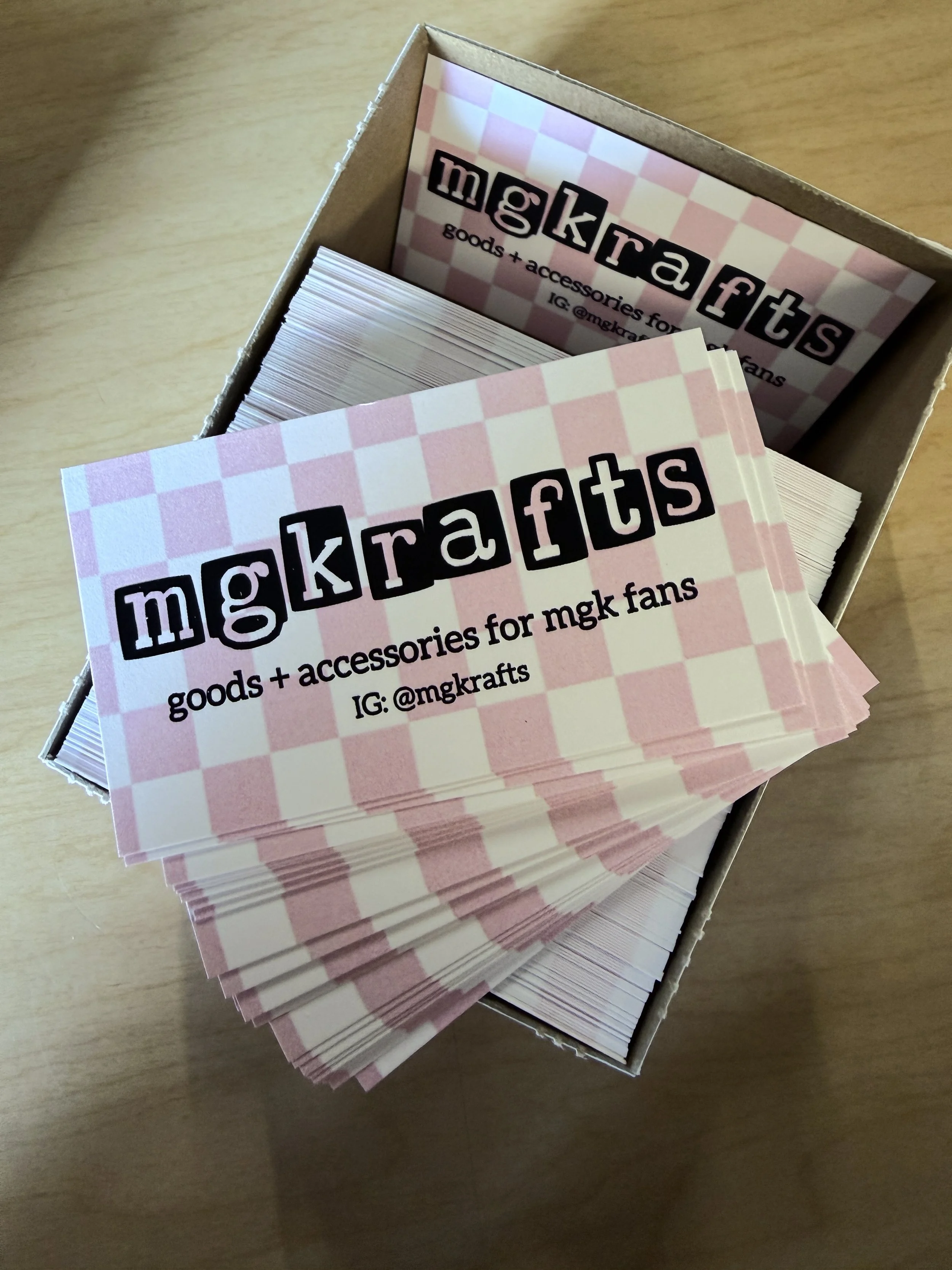 Stack of pink and white checkered business cards in a cardboard box, promoting MGKrafts accessories for MGK fans with Instagram handle @mgkrafters.